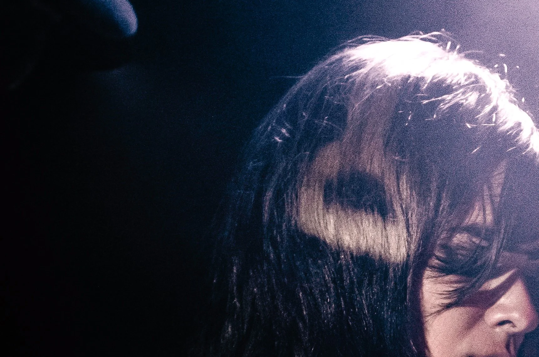 A woman with long, dark hair and glasses, looking slightly downward, illuminated by a light source from the right side of the image.