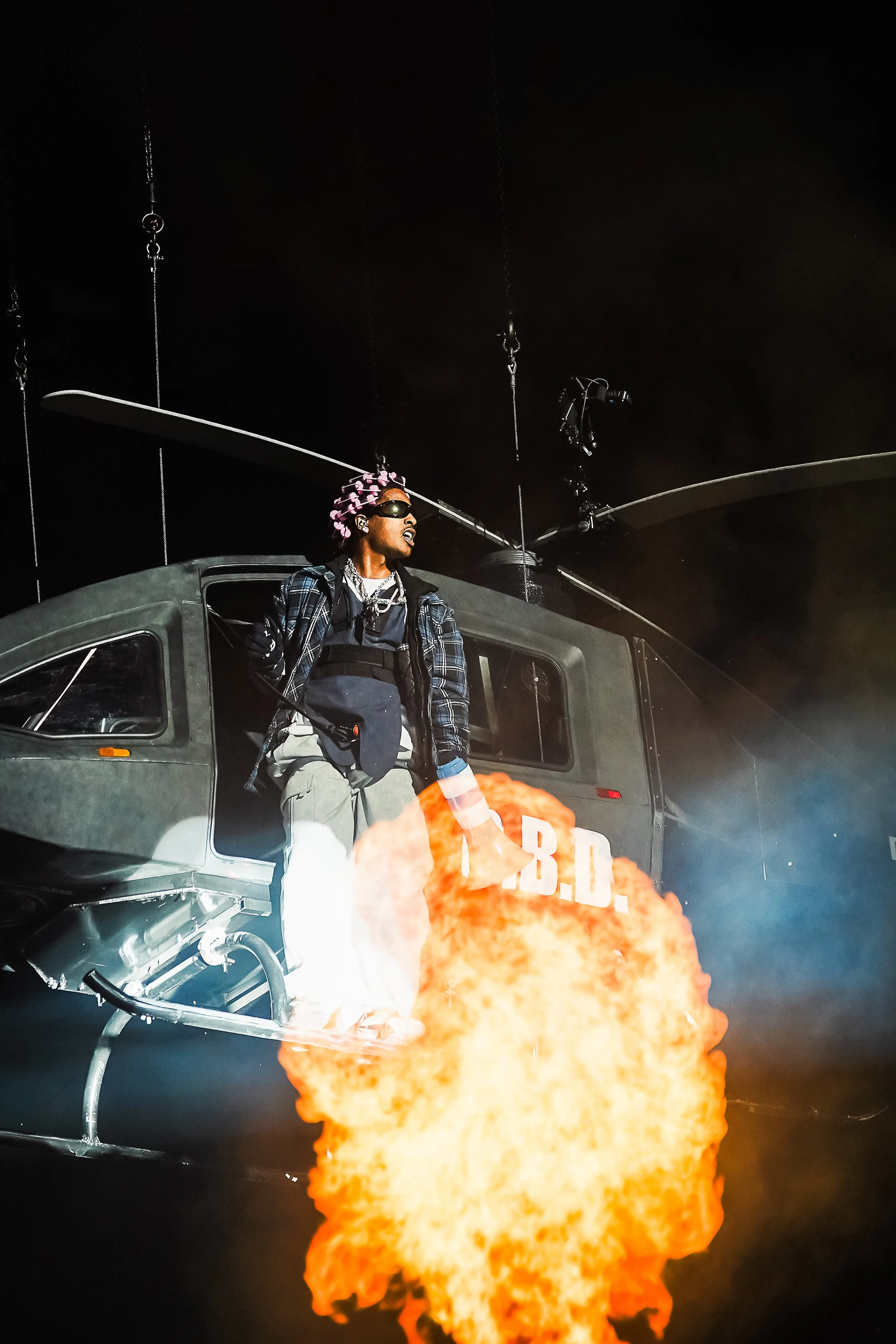 A person wearing sunglasses, a patterned headscarf, a plaid jacket, and layered clothing standing beside a helicopter at night, with flames or an explosion in the foreground.