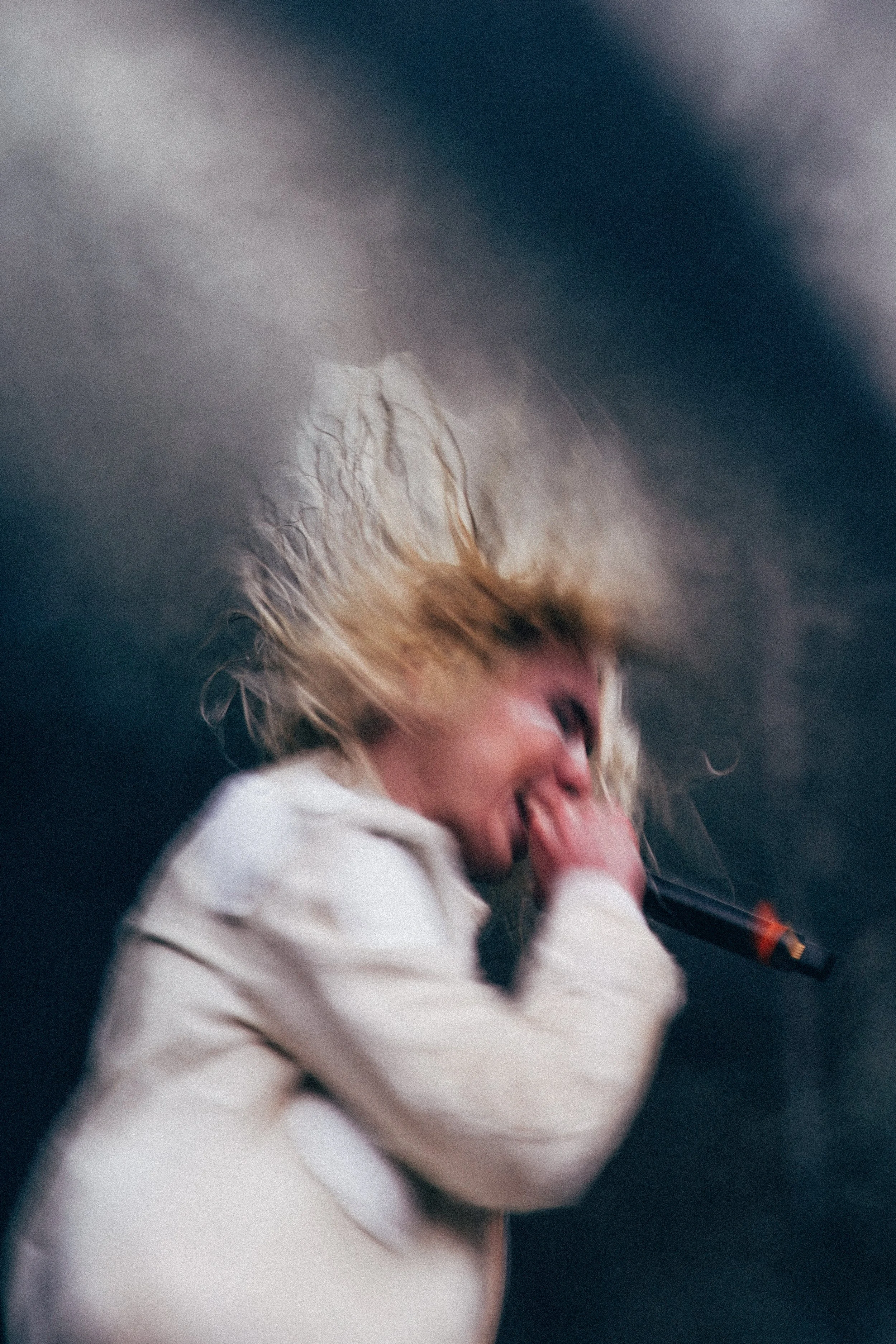 Person with blonde curly hair and light skin, wearing a white hoodie, smiling and holding a microphone.