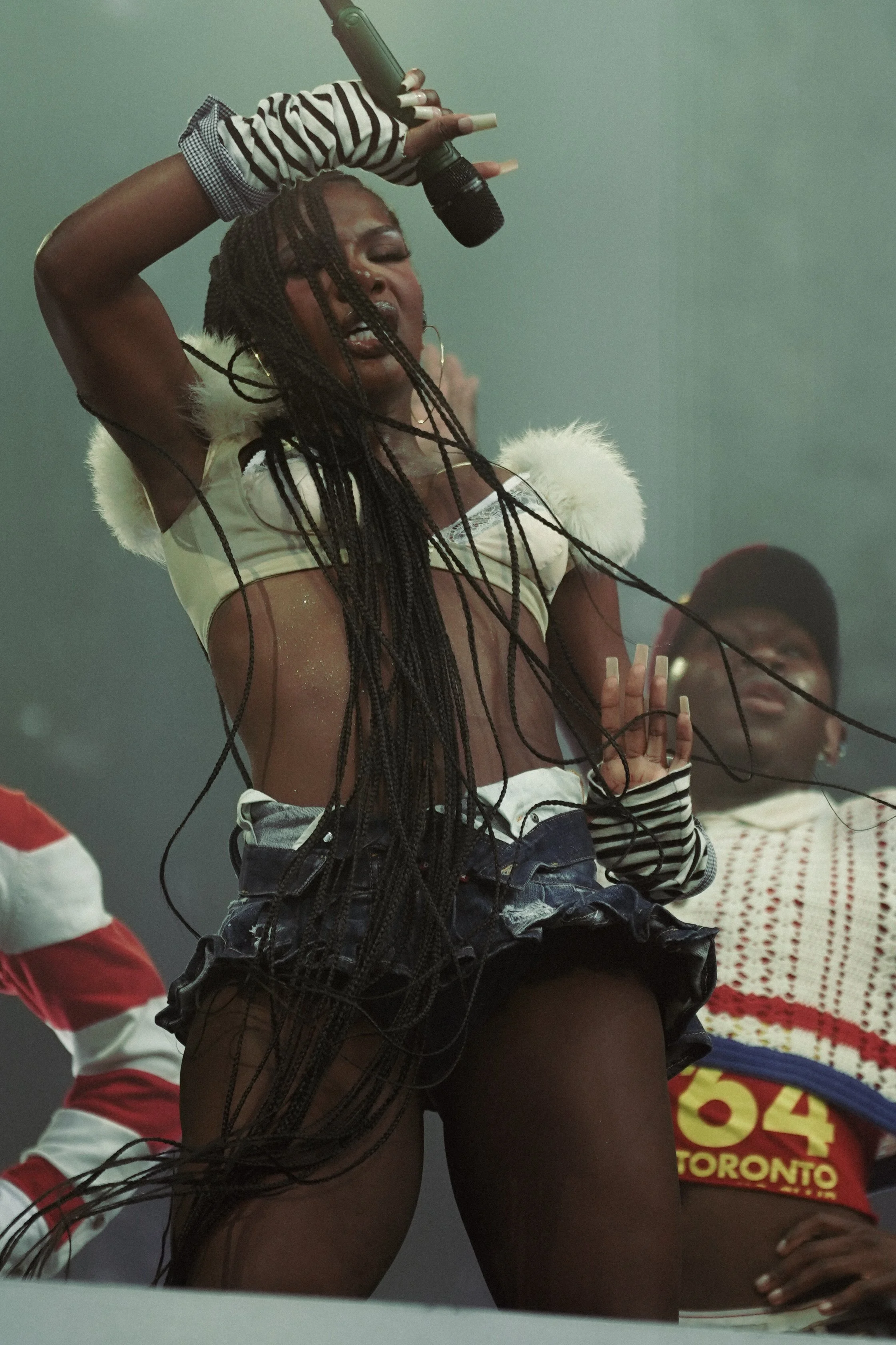A woman with long braided hair wearing a cropped top with fur-lined shoulders, black shorts, and striped gloves holds a microphone above her head, singing passionately. Others are in the background, one wearing a Toronto football jersey.