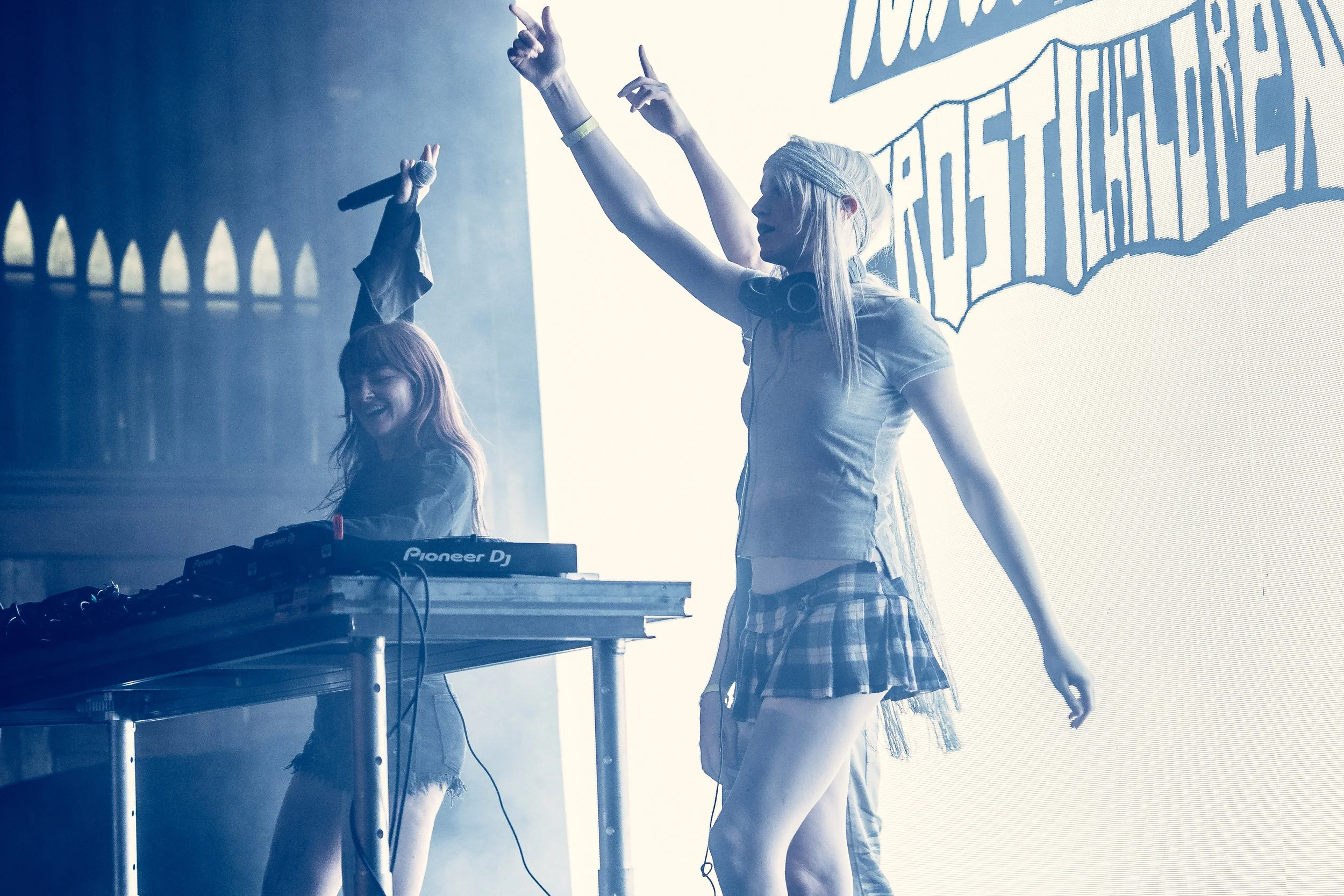 Two female DJs perform on stage with DJ equipment, one with headphones around her neck, in front of a large illuminated backdrop with text, at a lively music event.