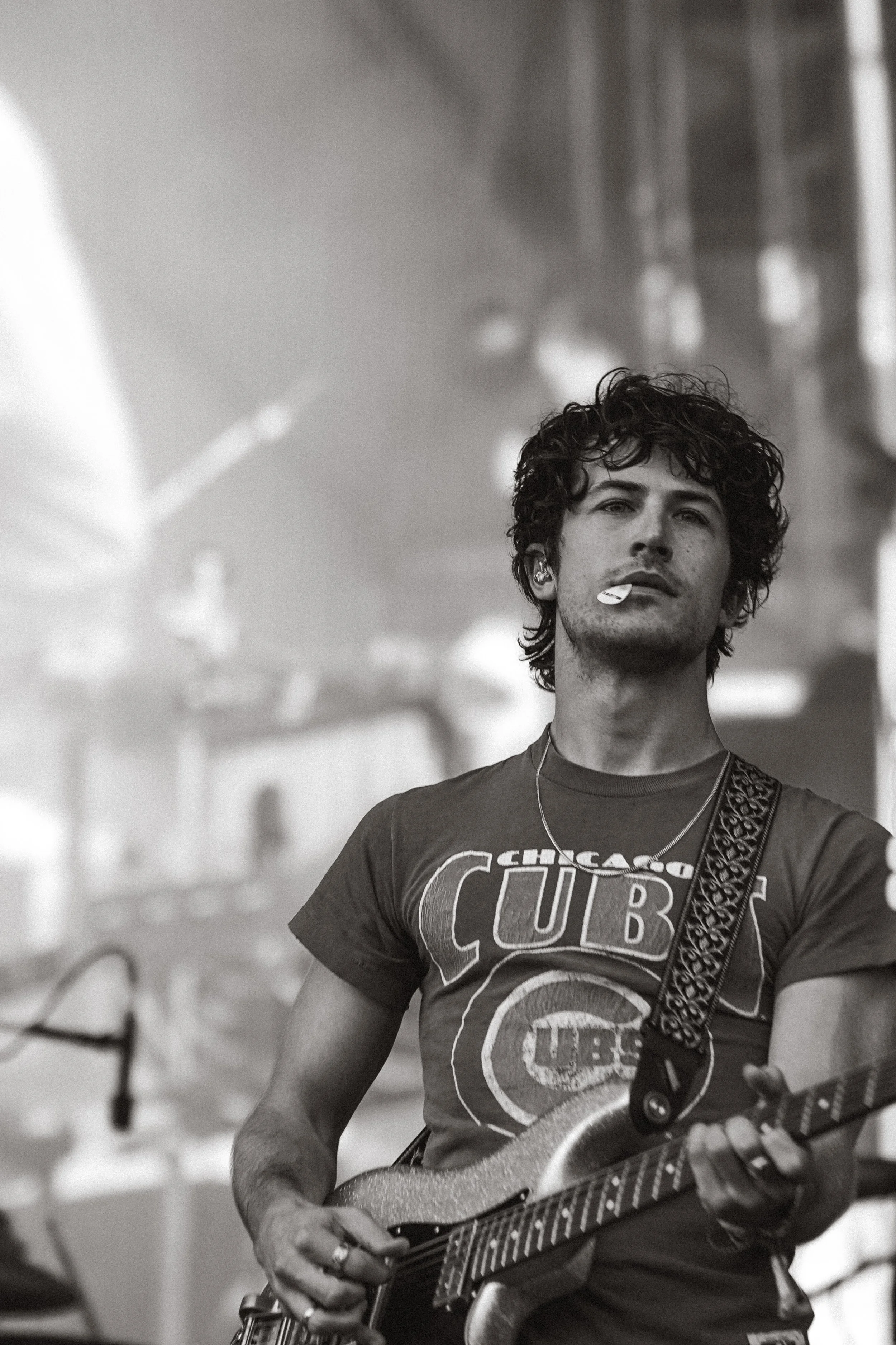 A man with curly hair wearing a Chicago Cubs T-shirt playing an electric guitar in a room with blurry background elements.