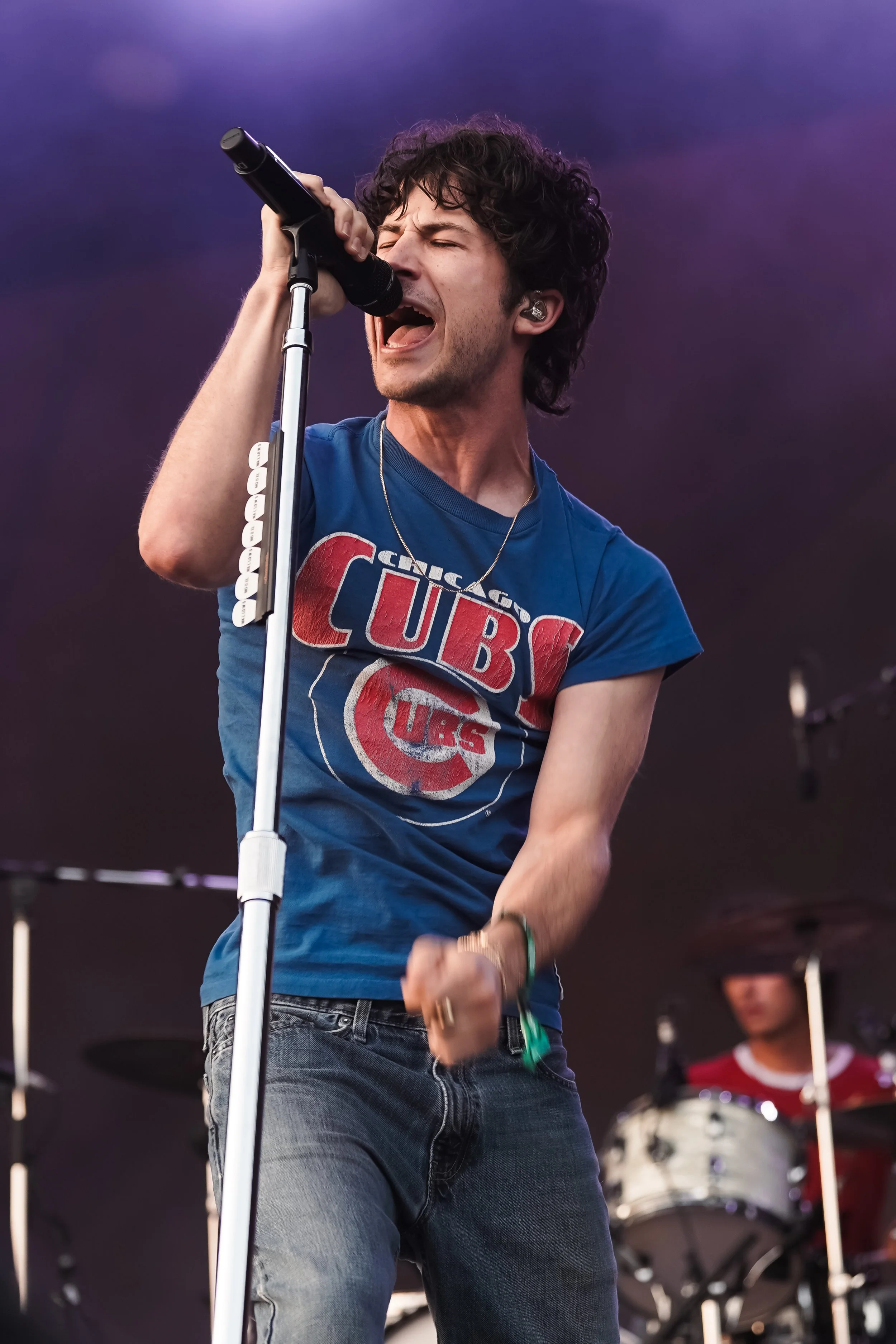 A male singer with curly hair passionately singing into a microphone during a live performance, with a drummer in the background.