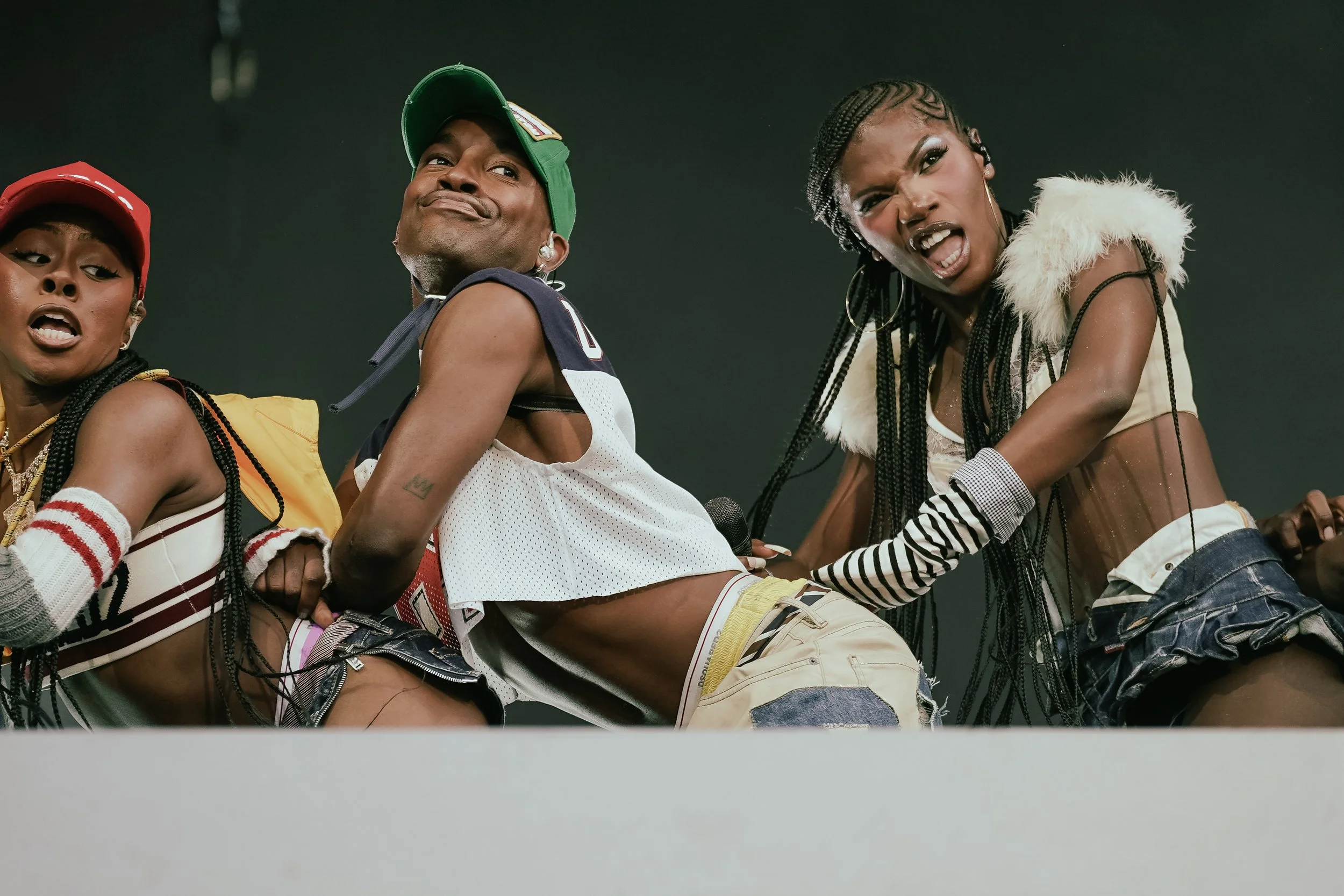 Group of four young women with expressive faces, dressed in trendy casual clothing, posing together against a dark background.