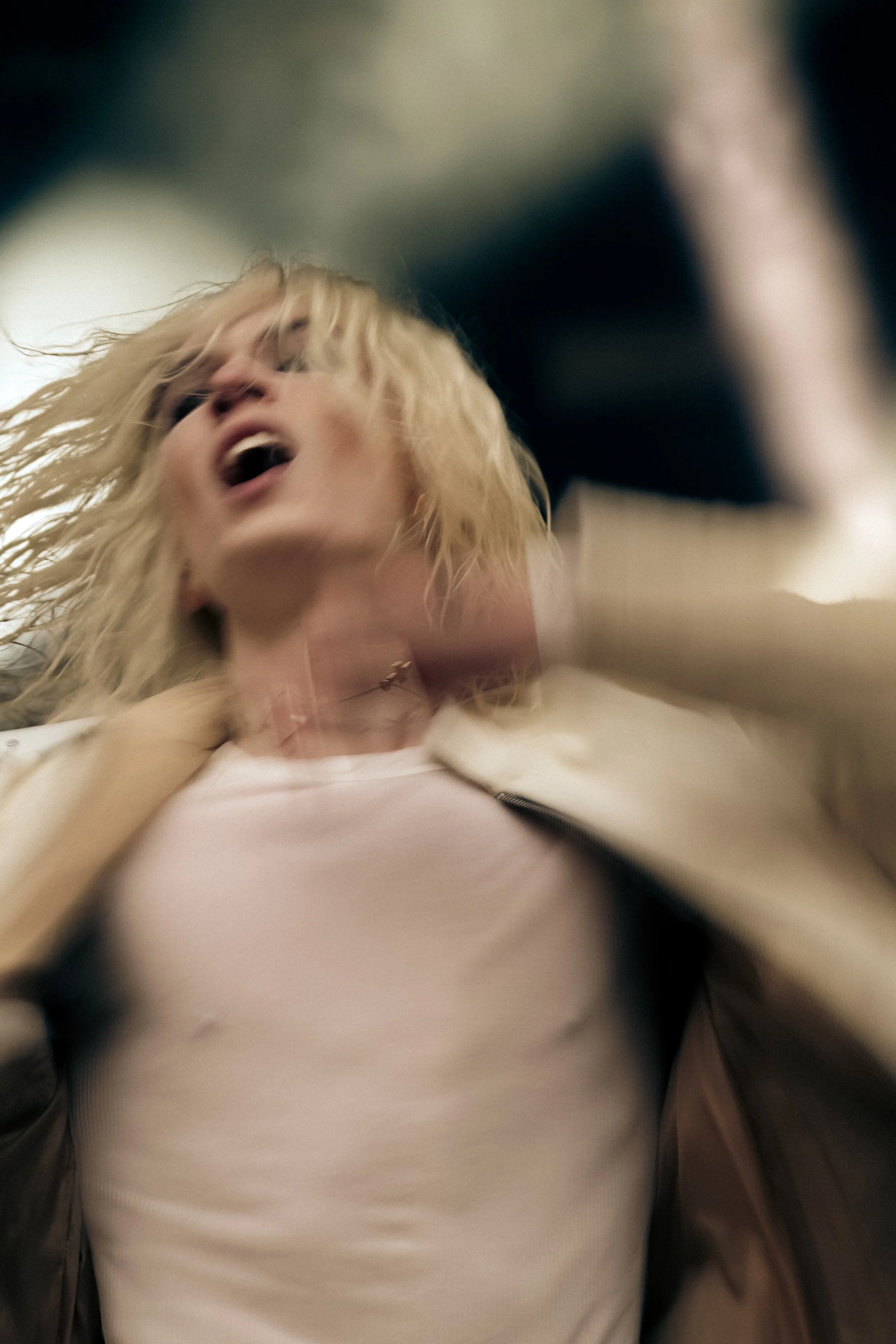 Close-up of a person with blonde hair, head tilted back, mouth open, and eyes closed, wearing a light-colored shirt and a jacket, against a blurred background.