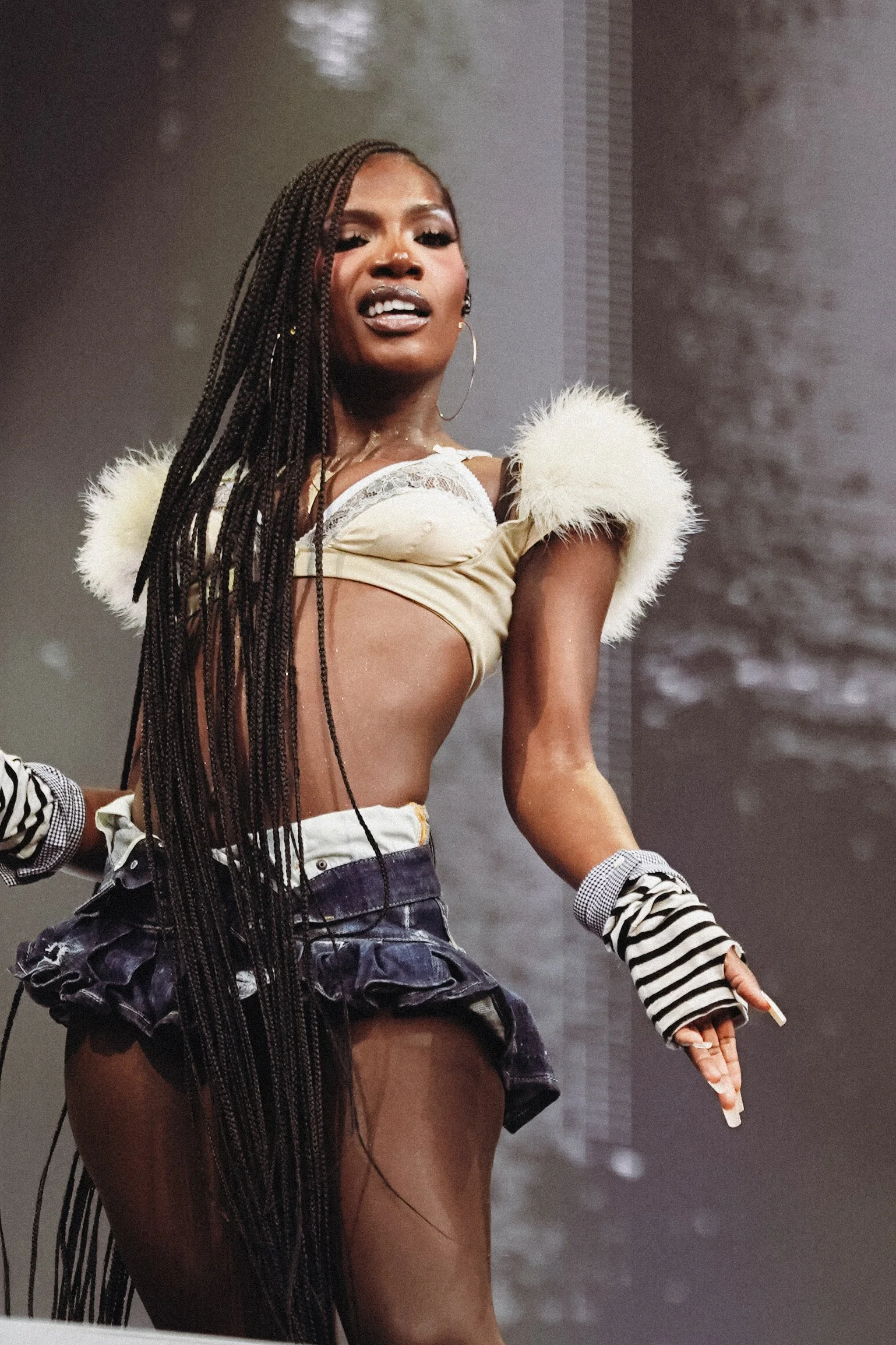 Close-up of a woman with long braided hair, wearing a cream-colored top with fluffy shoulder accents, large hoop earrings, and striped gloves, on stage.