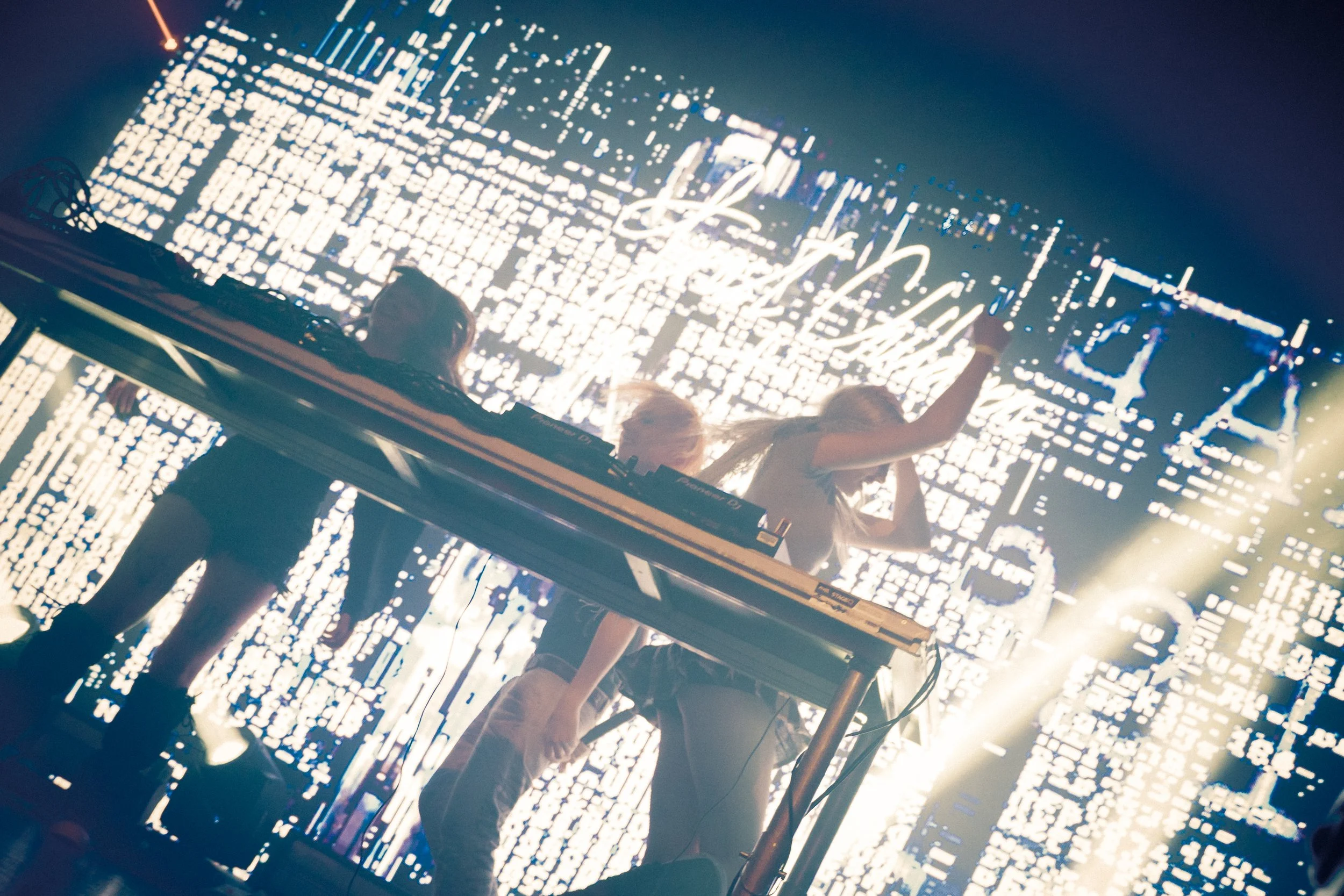 A DJ performs on stage with a LED light display behind her, holding a tablet in her right hand, with her left hand raised. The stage has a DJ booth and equipment, and there are people in the audience.