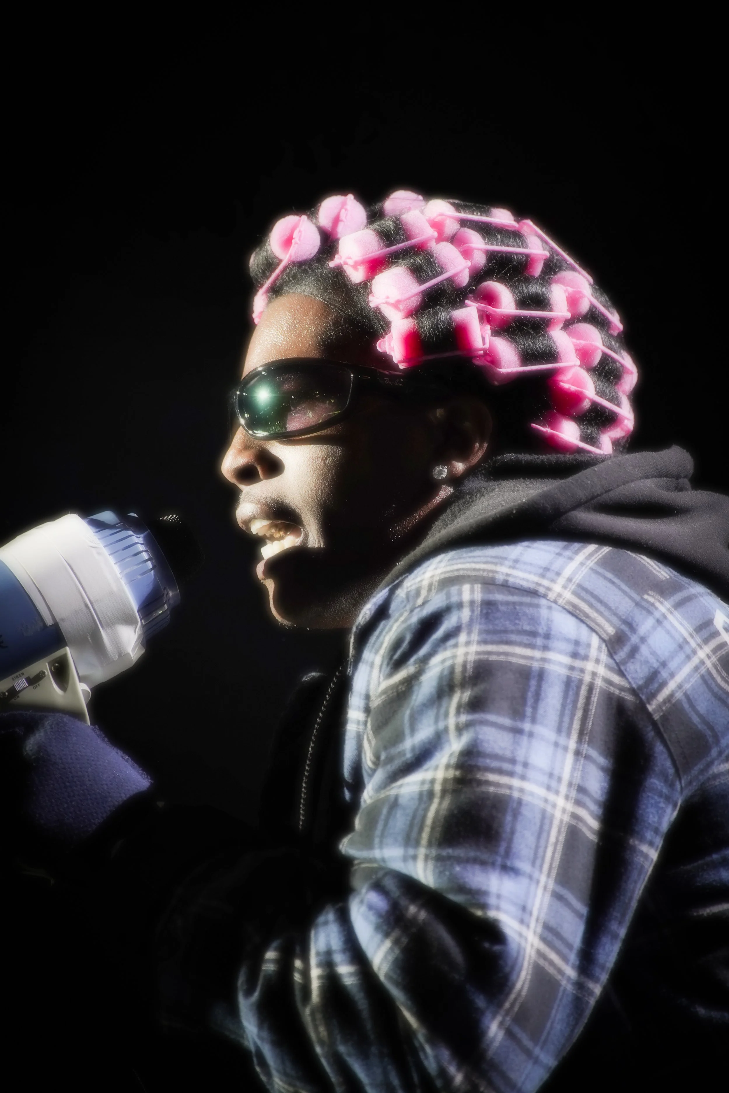 A woman wearing sunglasses and a plaid shirt is singing into a microphone. She has a black and pink hair roller set and appears to be performing passionately.