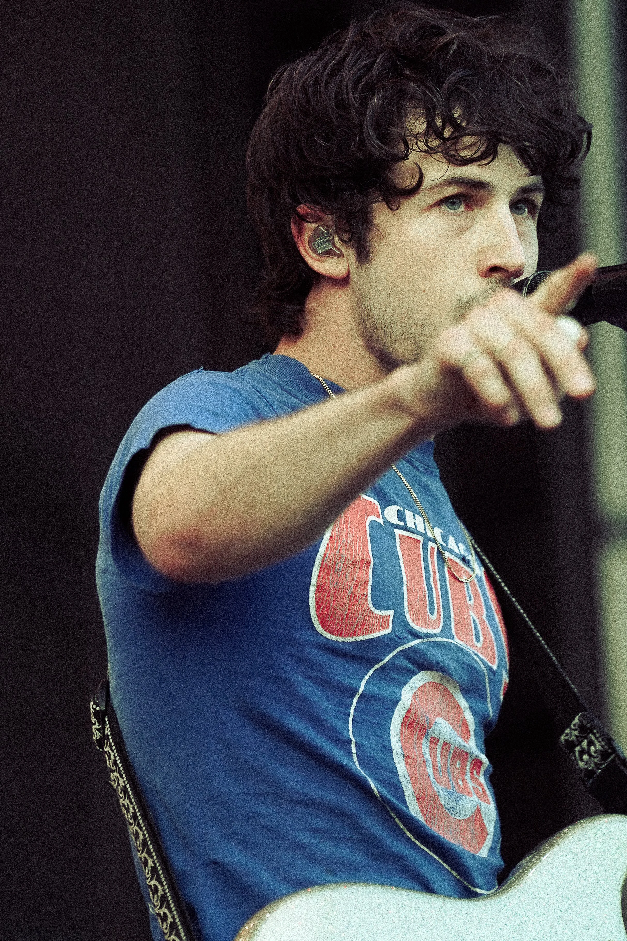 Close-up of a young man with curly dark hair, light skin, and blue eyes, wearing a blue T-shirt with a red, white, and blue logo. He is taking a sip from a straw or drinking device, with his hand visible and slightly blurred in the foreground. The ba