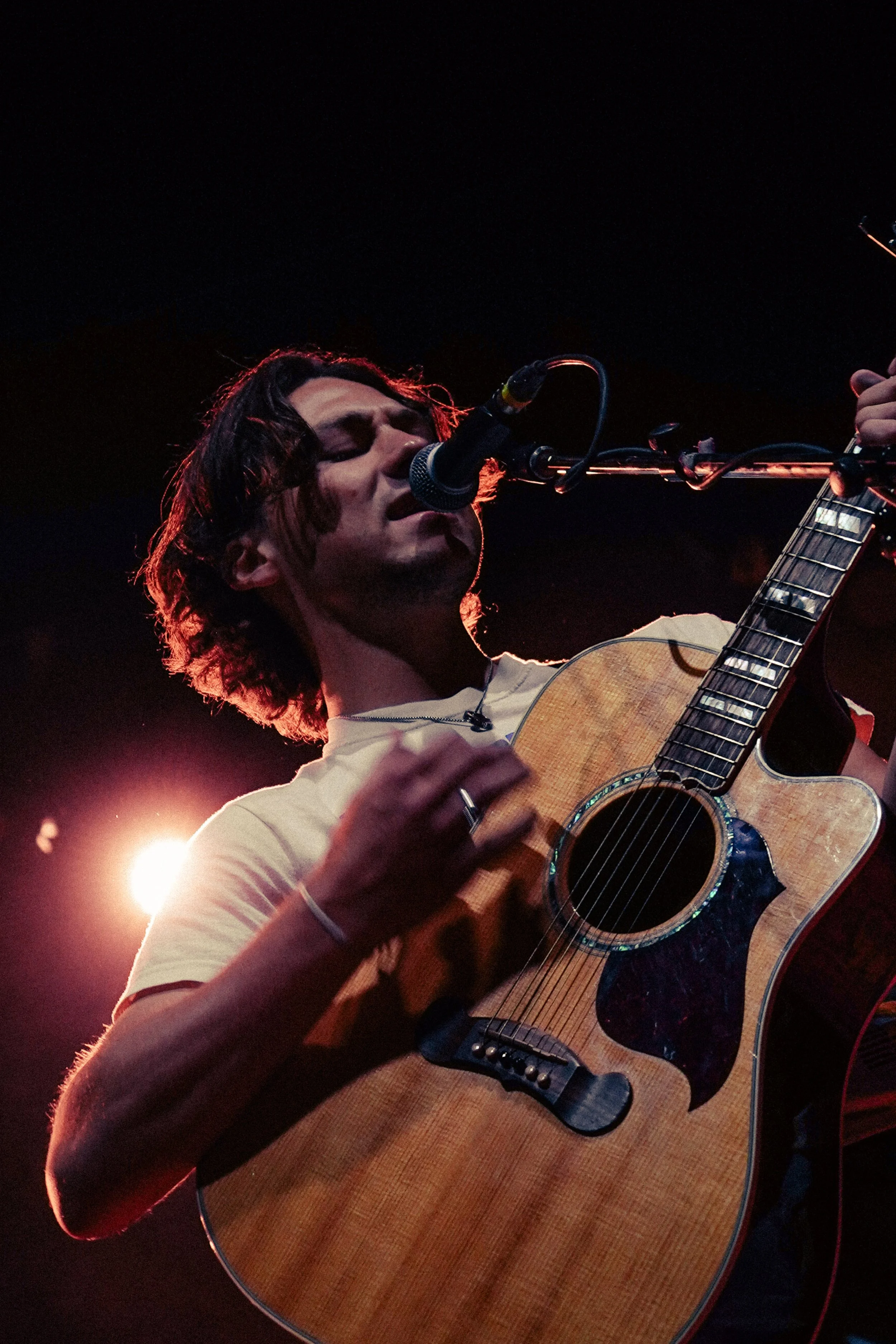 A male musician with long wavy hair performing on stage at night, playing an acoustic guitar and singing into a microphone, illuminated by stage lights.