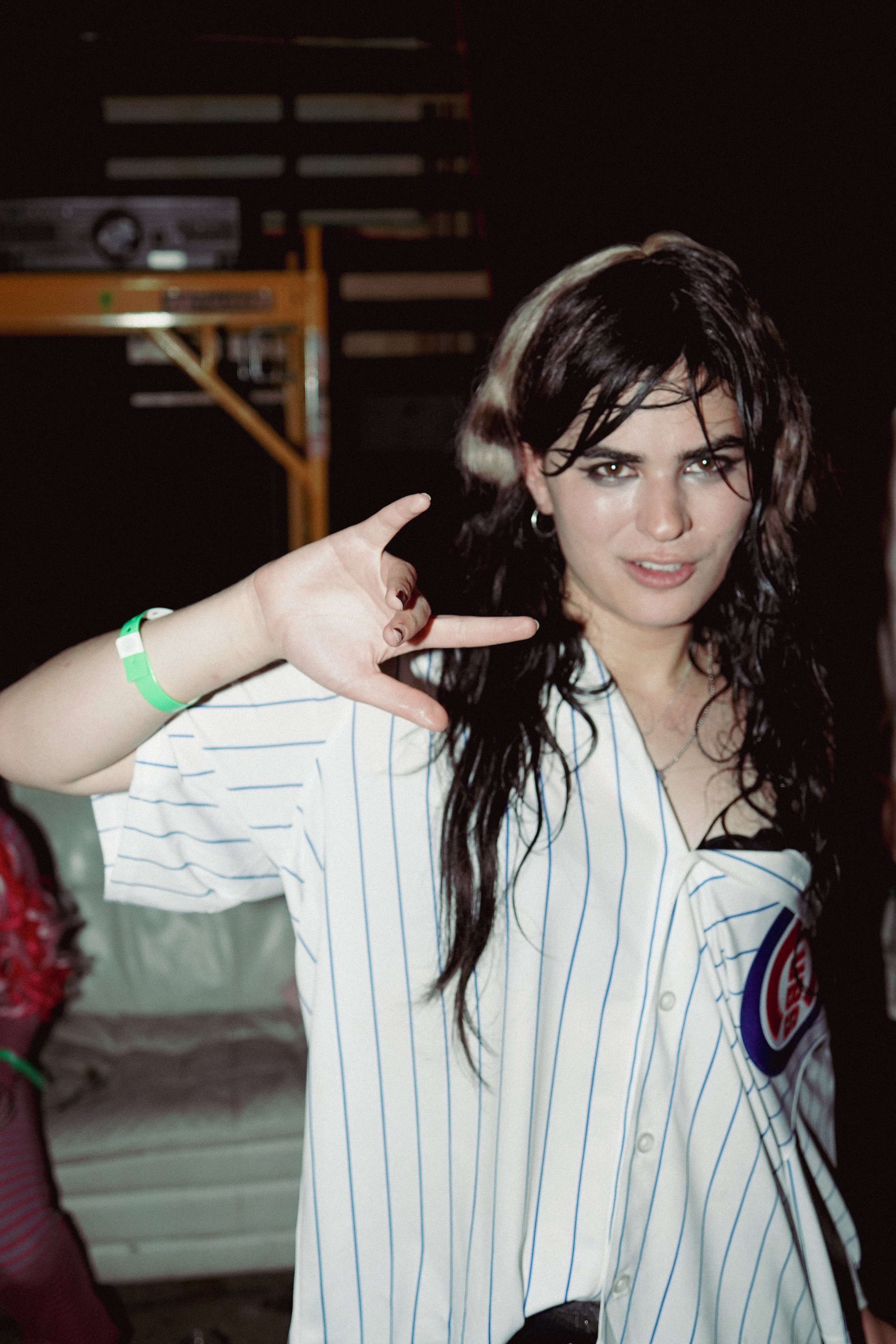 A woman with long dark wavy hair wearing a white baseball jersey with blue pinstripes and a logo on the sleeve making a peace sign with her right hand, standing indoors.
