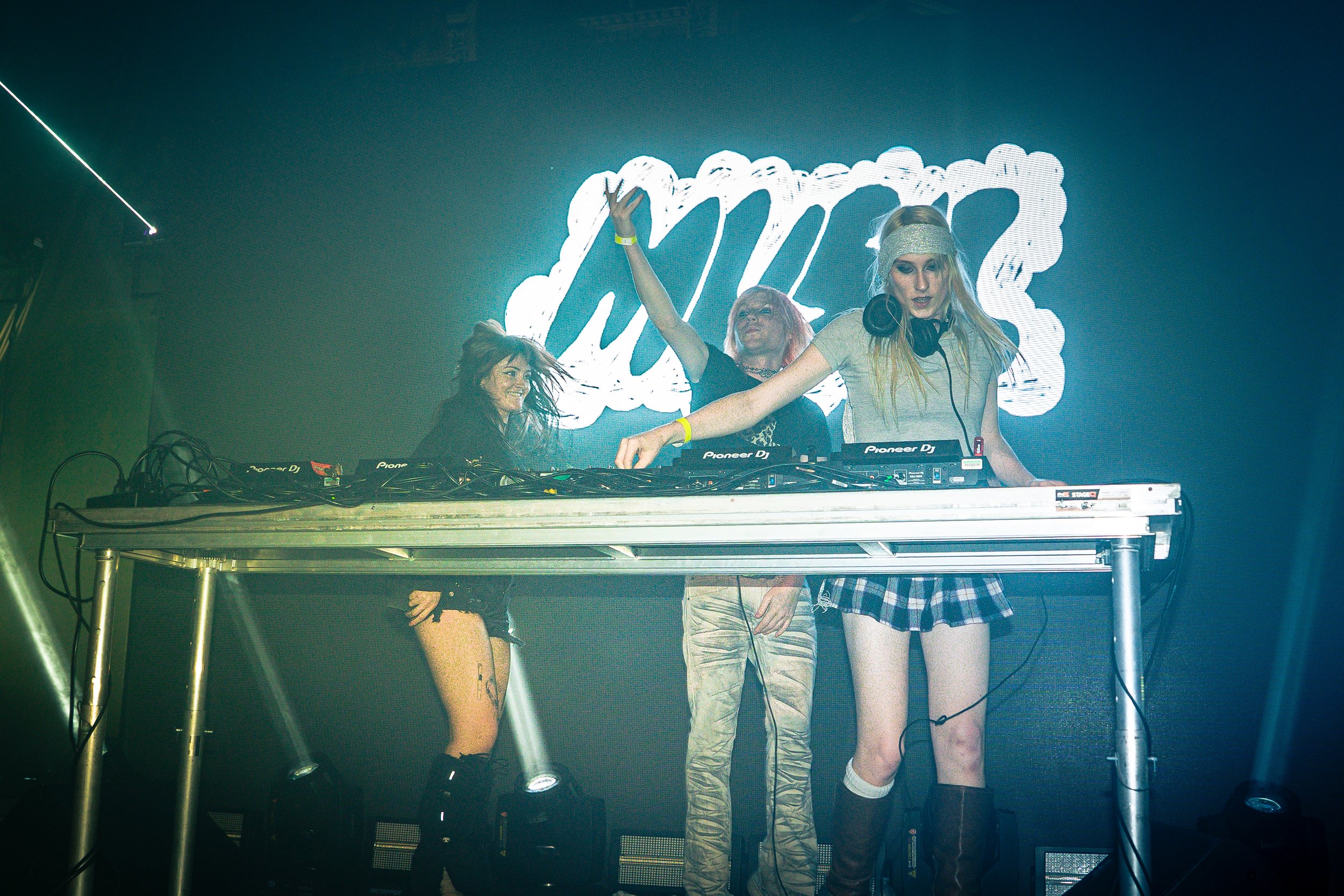 A female DJ wearing headphones and a headband, performing on stage with two women dancing nearby at a music event or party.