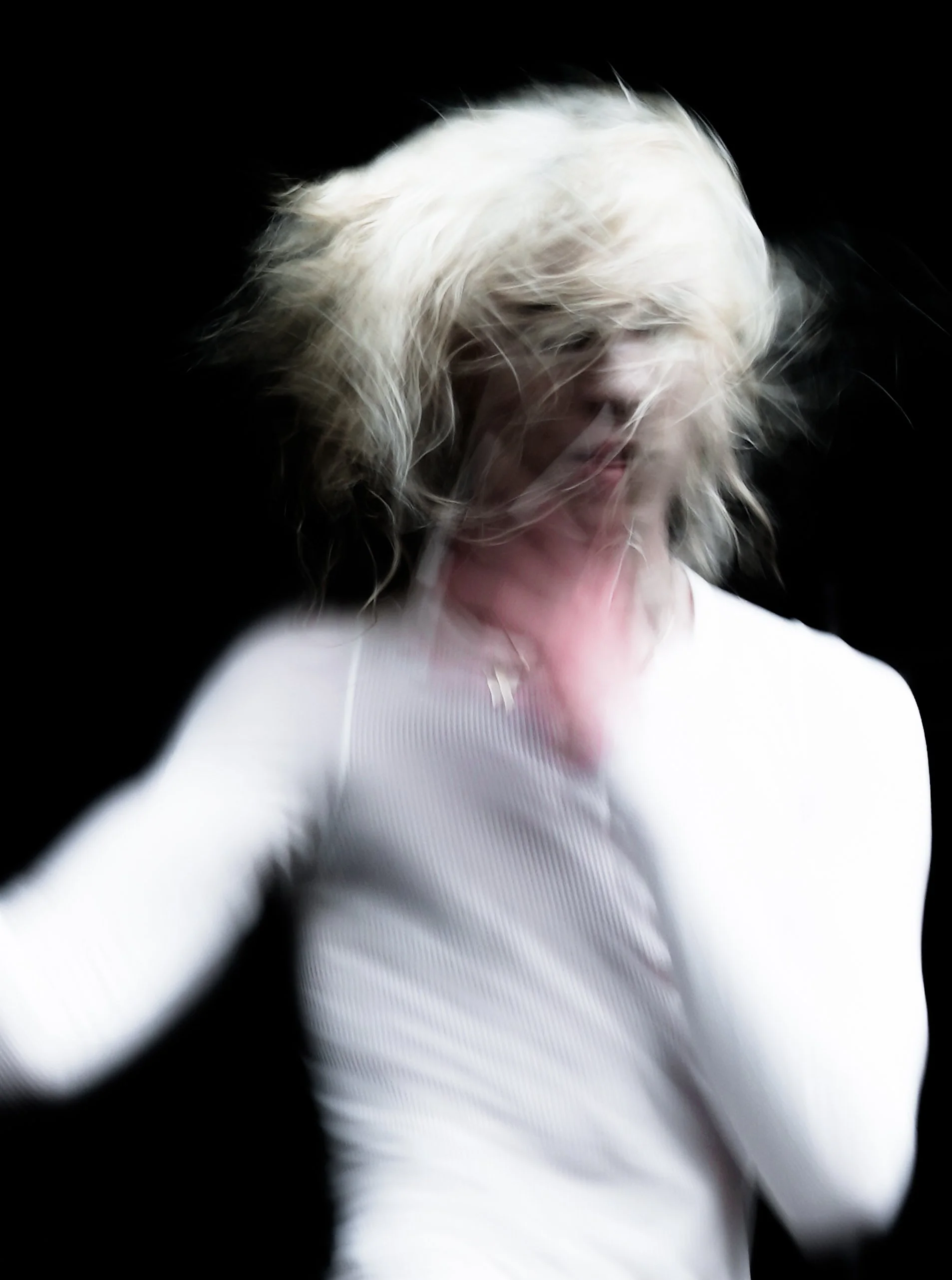 A person with light blonde hair wearing a white shirt, slightly bowed head, and hair covering their face, against a black background.