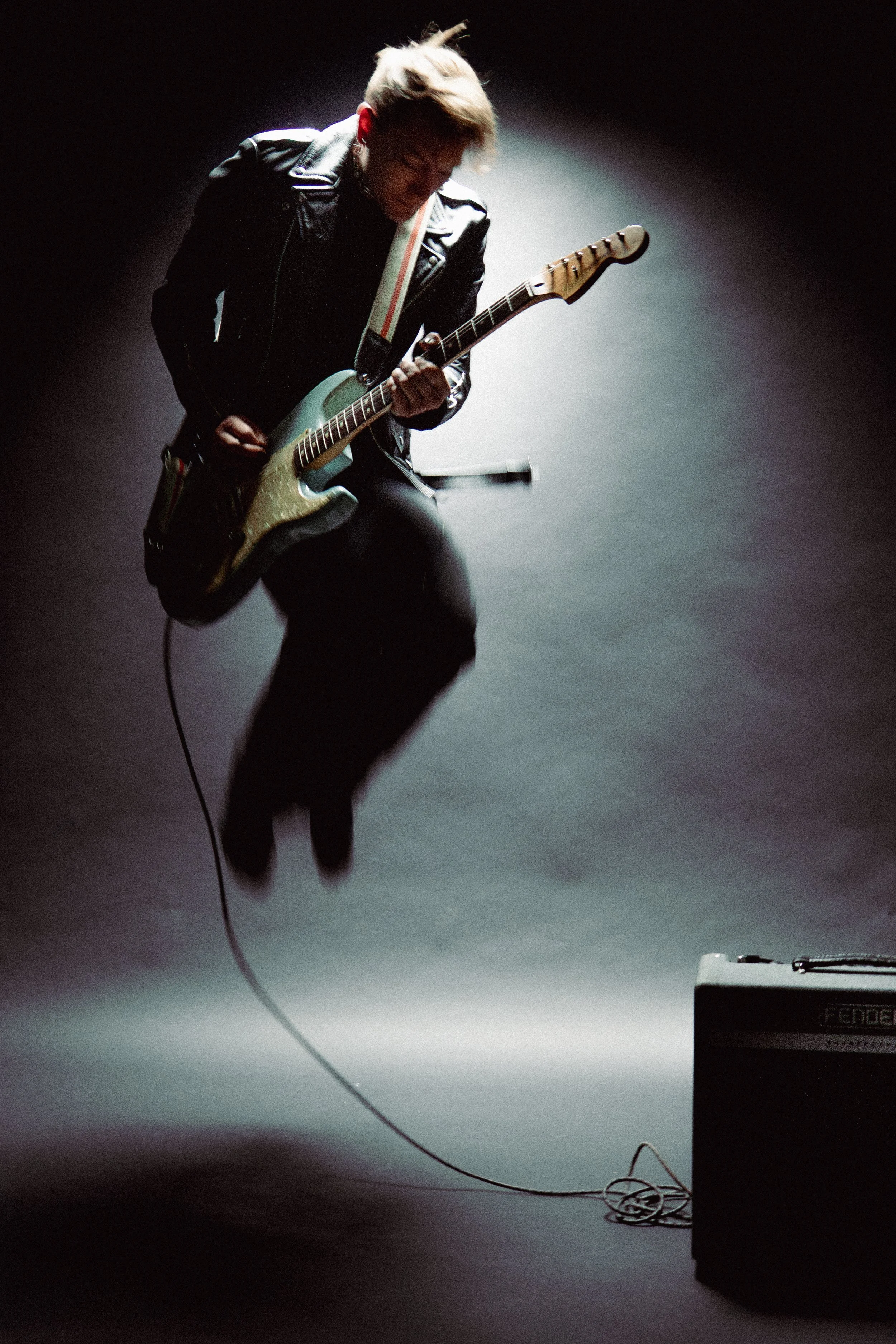 A person playing an electric guitar on stage, wearing a leather jacket, with a spotlight highlighting them against a dark background.