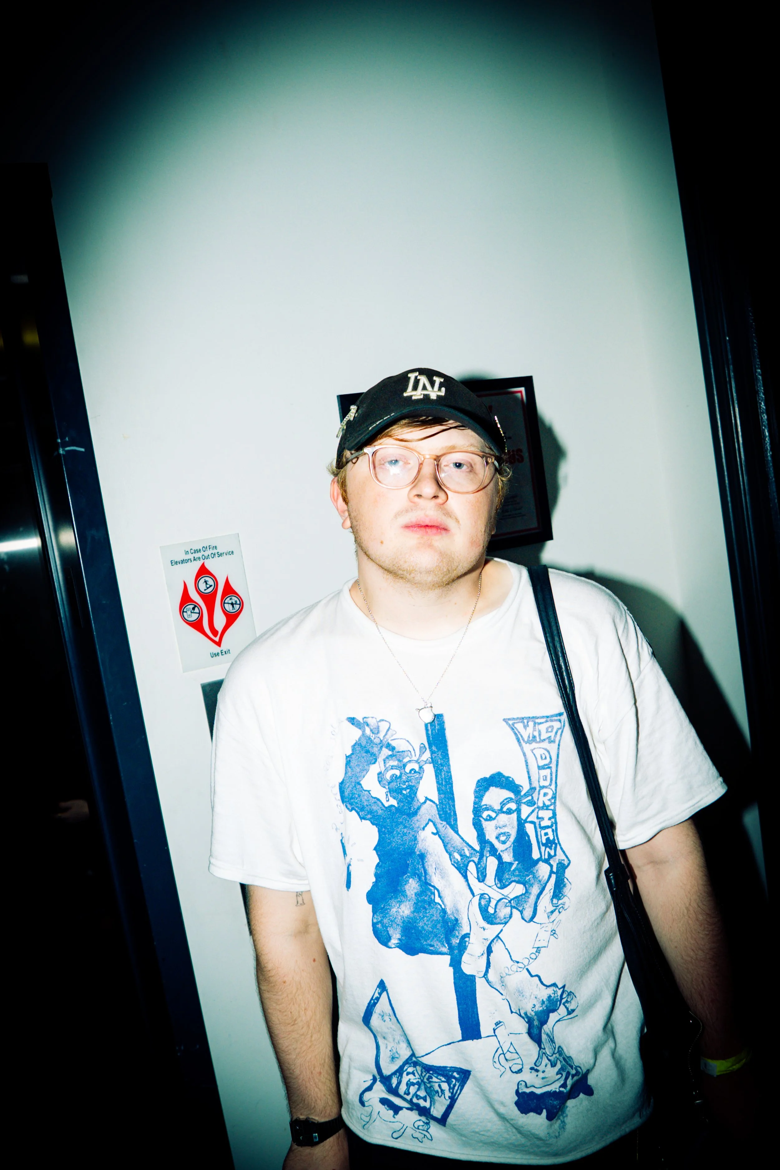 A young man with light skin, wearing glasses, a black cap with 'LA' on it, a white graphic T-shirt featuring cartoon characters, and a black watch. He is standing in front of a white wall with a framed award and a safety sign. His expression is neutr