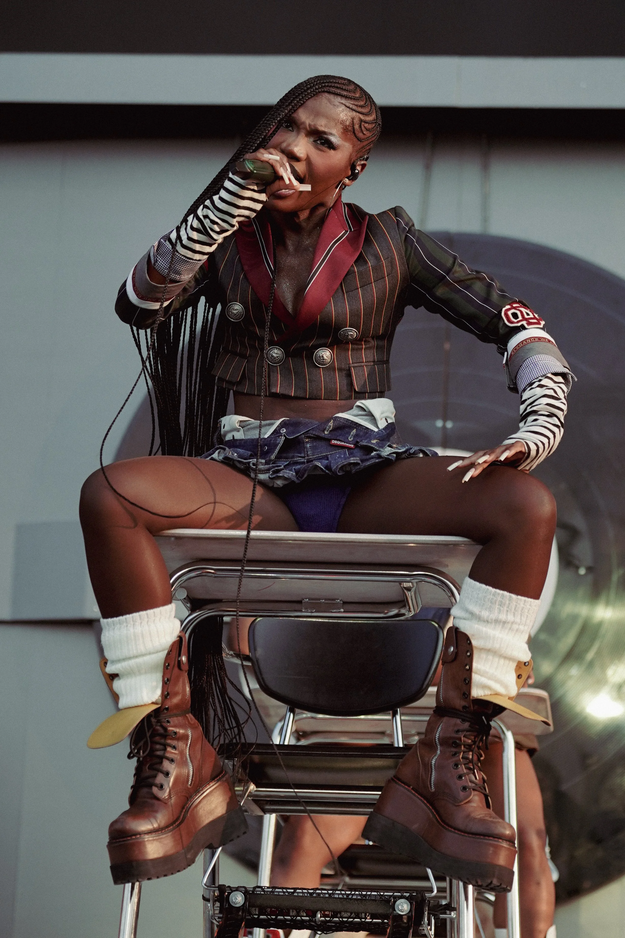 A woman with braids, wearing a striped crop top, blazer, shorts, and brown boots, sitting on a chair and holding a cigarette, outdoors in front of a modern building.