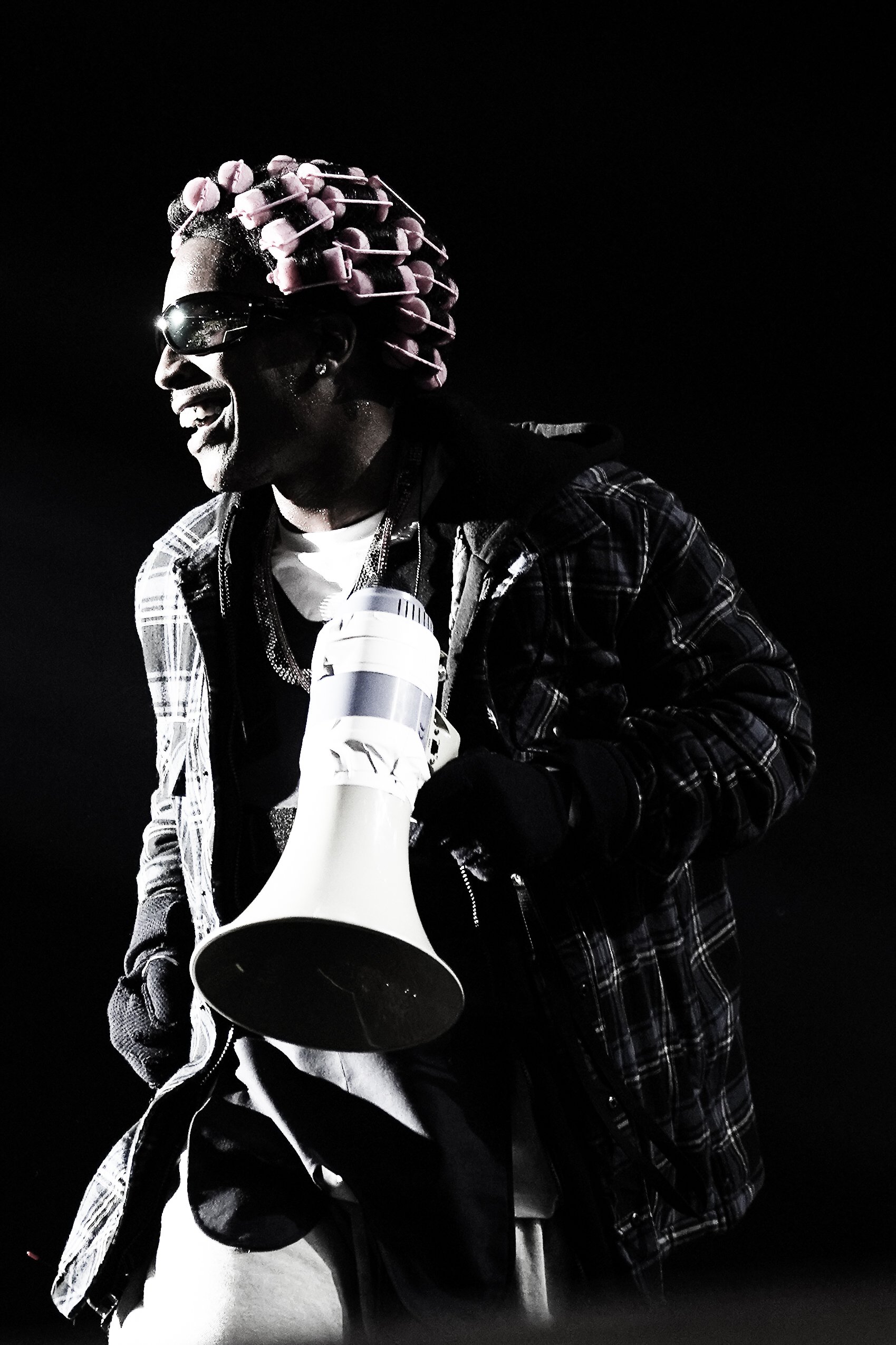 A person wearing sunglasses and a plaid jacket is smiling and holding a large megaphone. They have pink curlers in their hair and are wearing a necklace, with a black background.