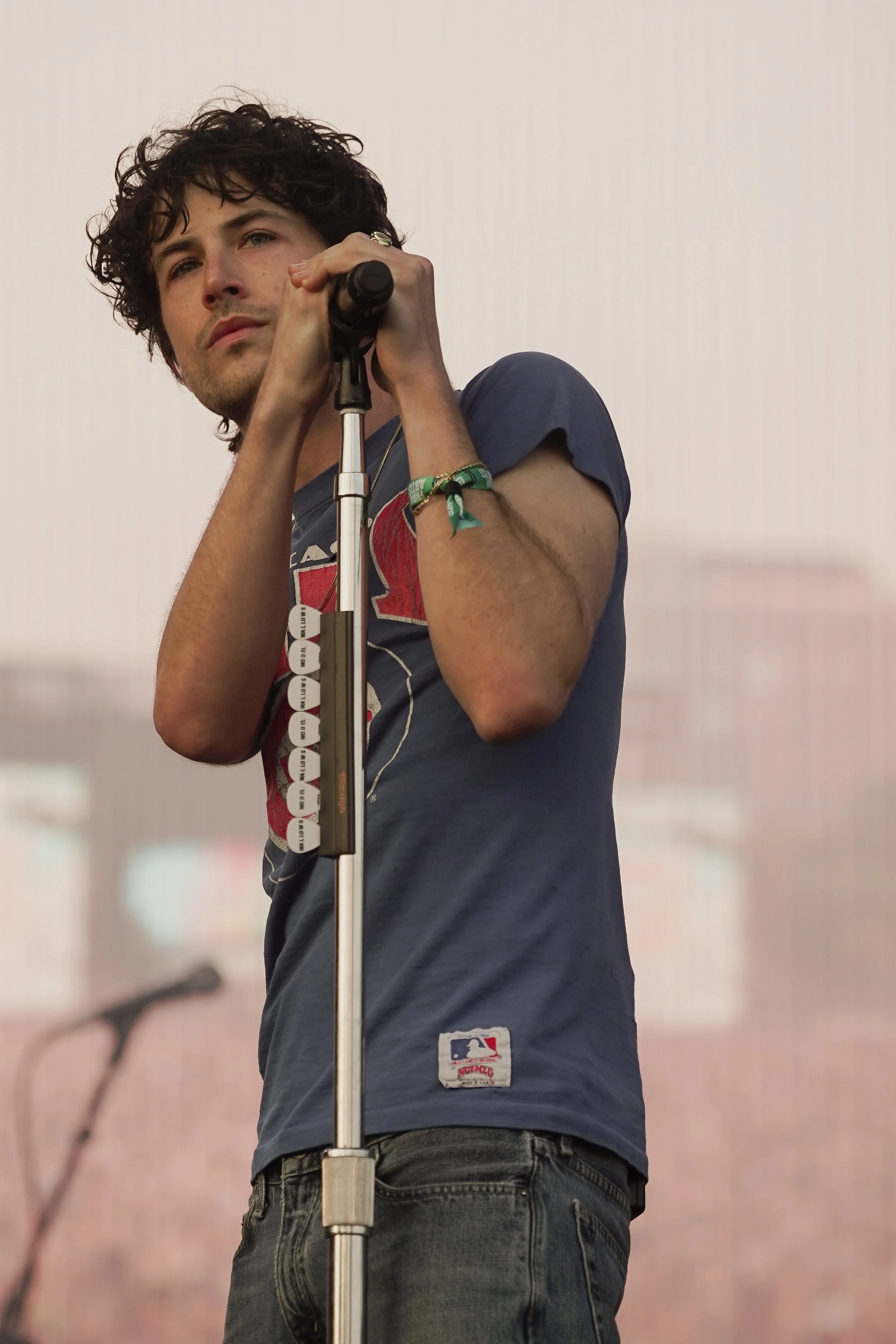 A man with curly dark hair and light skin, wearing a blue T-shirt with a sports logo, standing next to a microphone stand in an indoor setting with a plain, light-colored background.
