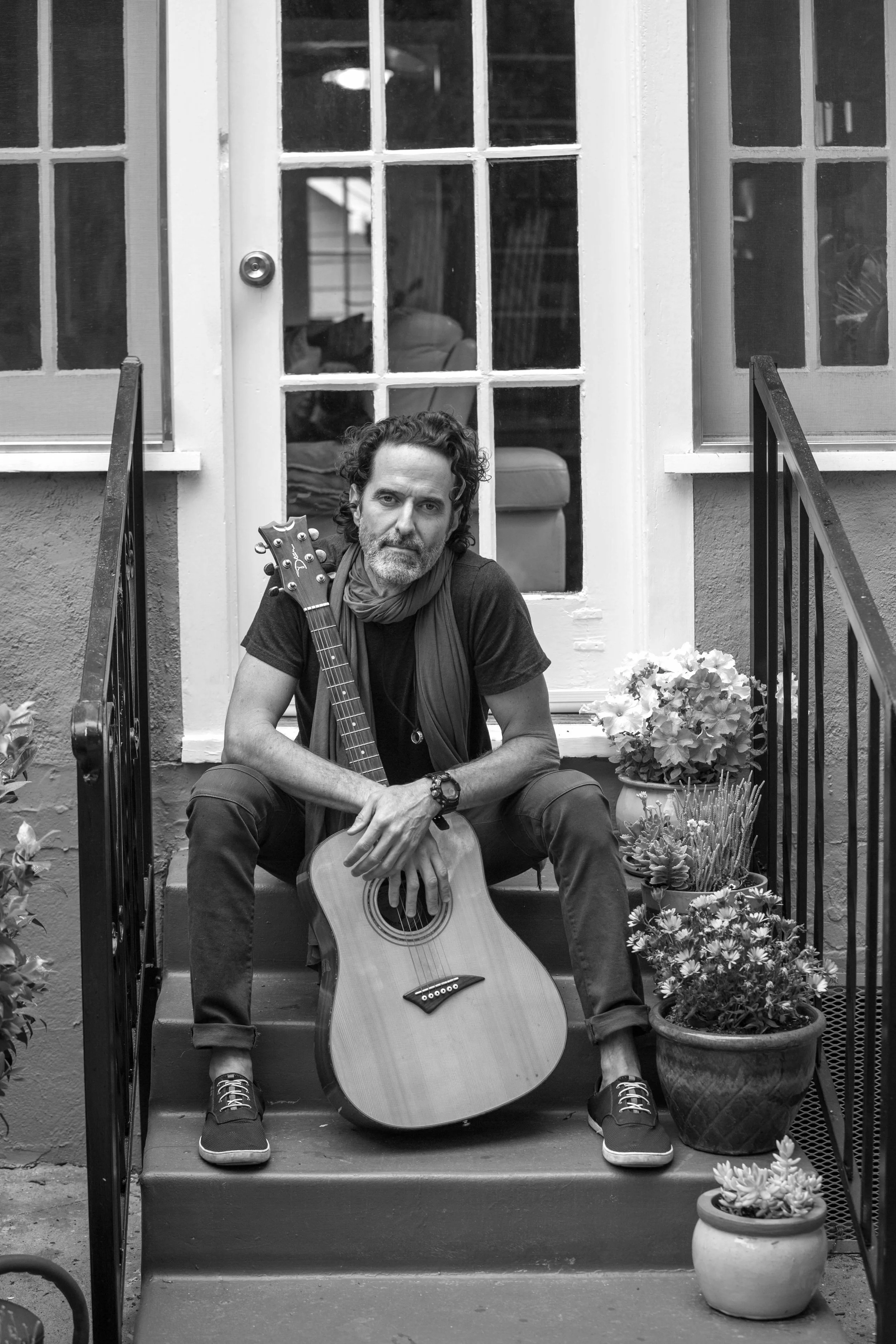 A man with curly hair, a beard, and a scarf sits on steps holding an acoustic guitar. There are potted plants on the steps and behind a glass door. The image is in black and white.