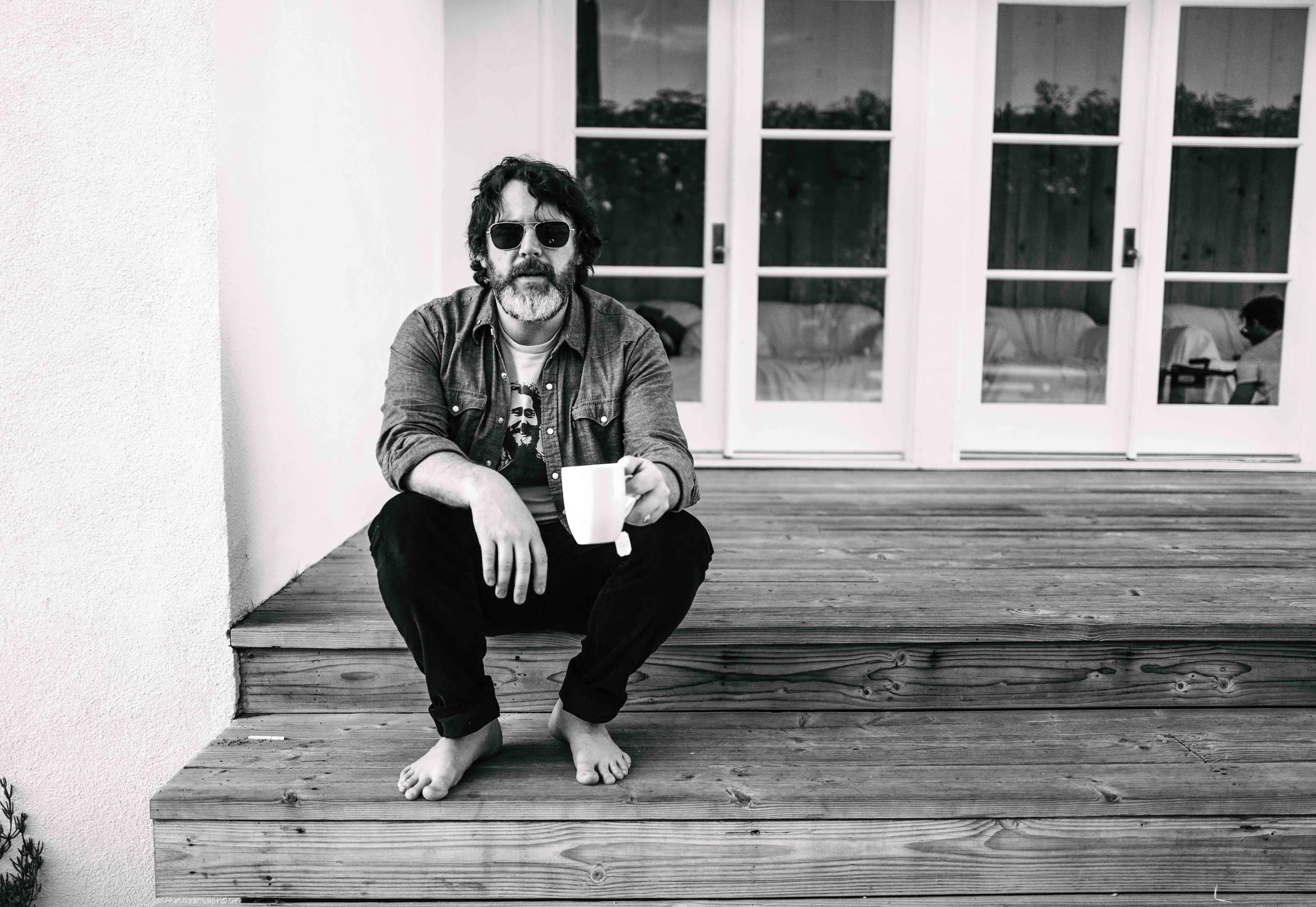 A man with dark curly hair and a beard, wearing sunglasses, a graphic t-shirt, and a denim jacket, sits barefoot on a wooden deck holding a white mug. Behind him are large glass doors with reflections, and he is leaning against a white wall.