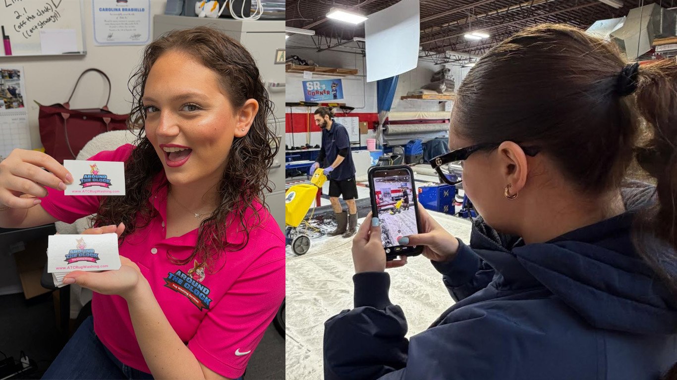 A woman in a pink shirt smiling and holding business cards with a logo, while another person takes a photo of her with a smartphone in a workshop or industrial setting.