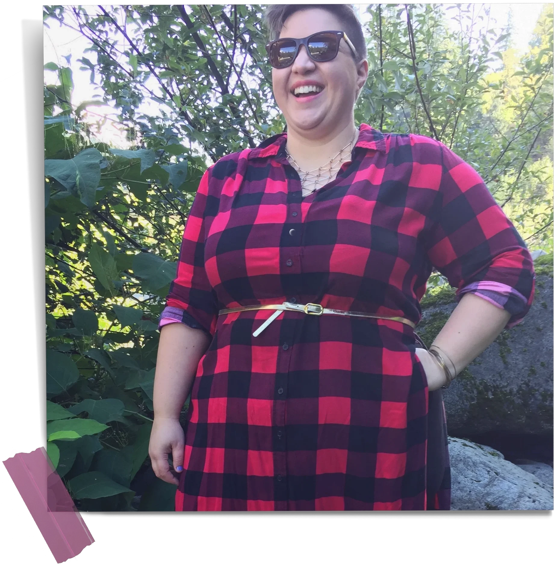 Laura with short hair wearing black sunglasses, a red and black plaid dress, a geometric gold necklace, and a gold belt stands grinning outdoors in Leavenworth, Washington.