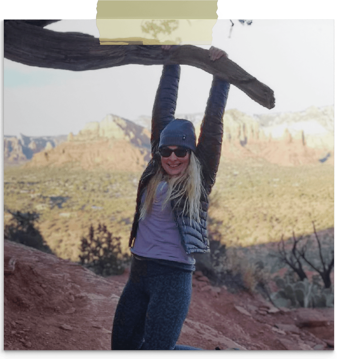Lizzie with long blond hair, wearing sunglasses, a beanie, a black jacket, and gray leggings, smiling and holding onto a tree branch in a desert landscape with canyon cliffs in the background.