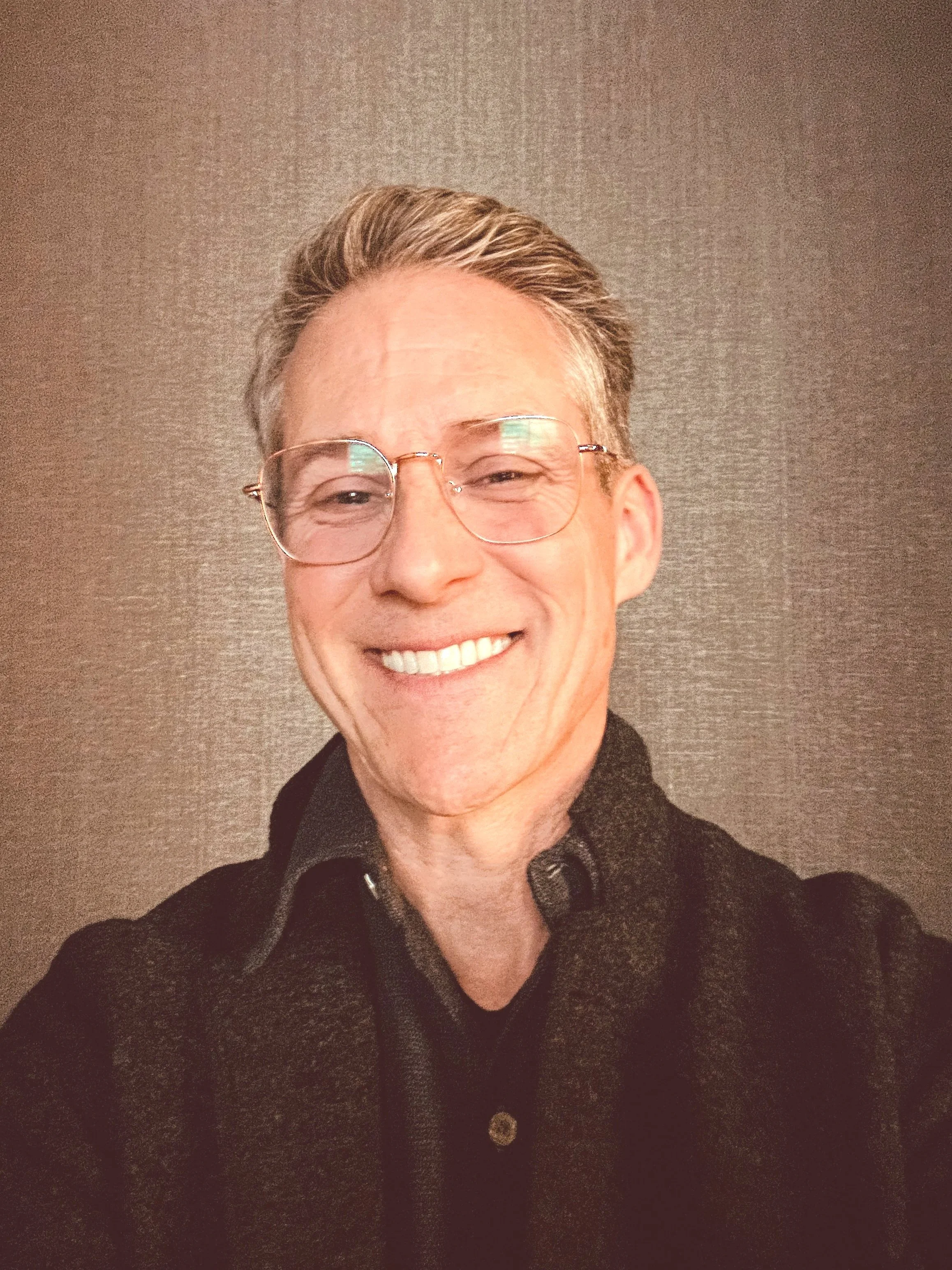 Close-up of a smiling man with glasses and gray hair, wearing a black shirt, standing against a textured gray wall.