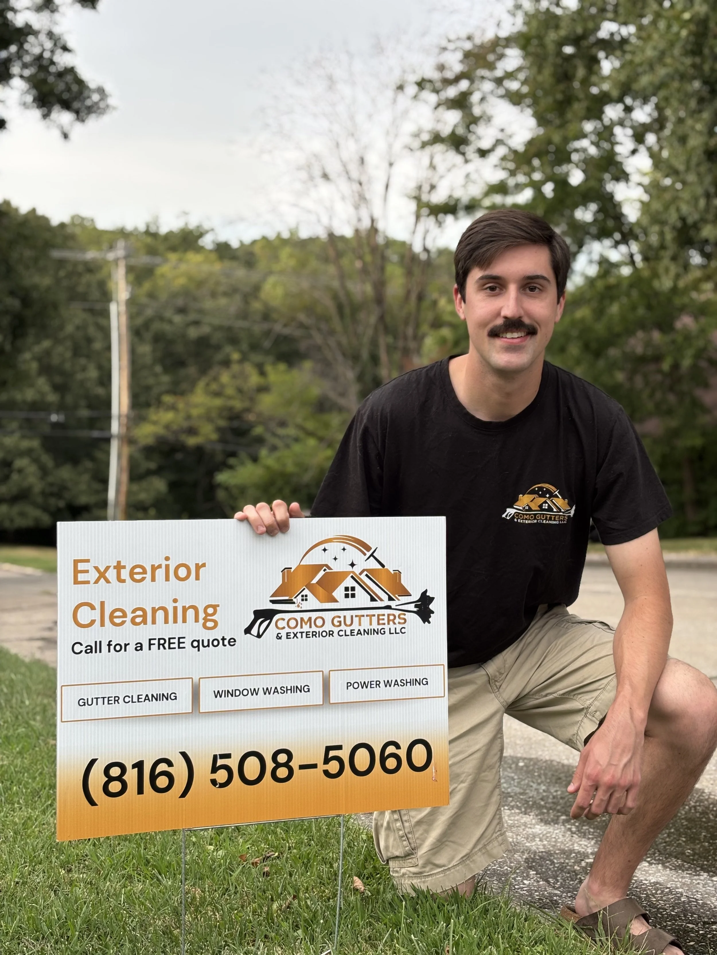 A man crouching outdoors holding a sign for Como Gutters & Exterior Cleaning LLC, advertising exterior cleaning services. He is wearing a black T-shirt with the company logo and khaki shorts. The background shows trees and a cloudy sky.