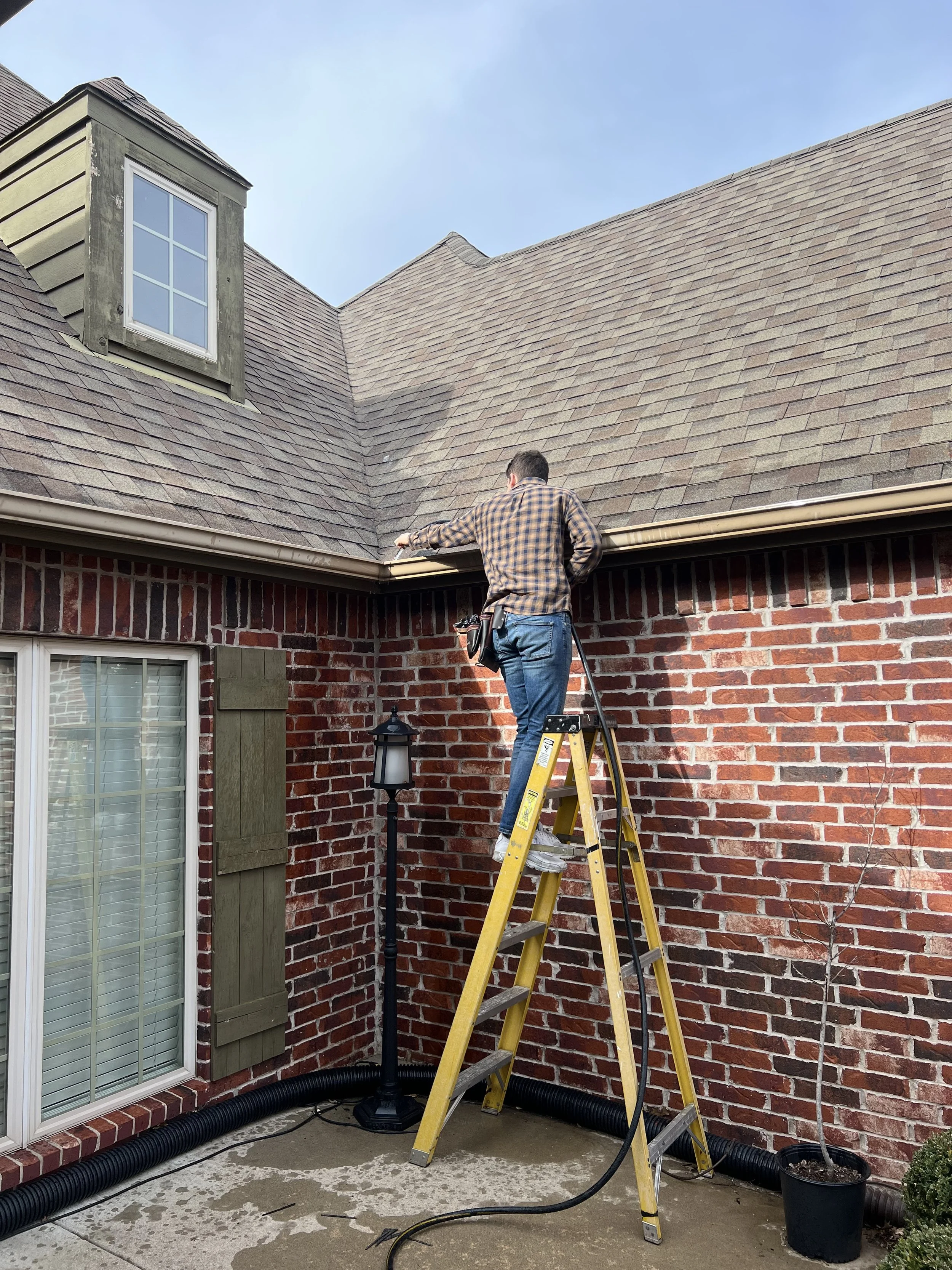 A man in a checkered shirt standing on a yellow ladder, working on the roof of a house.