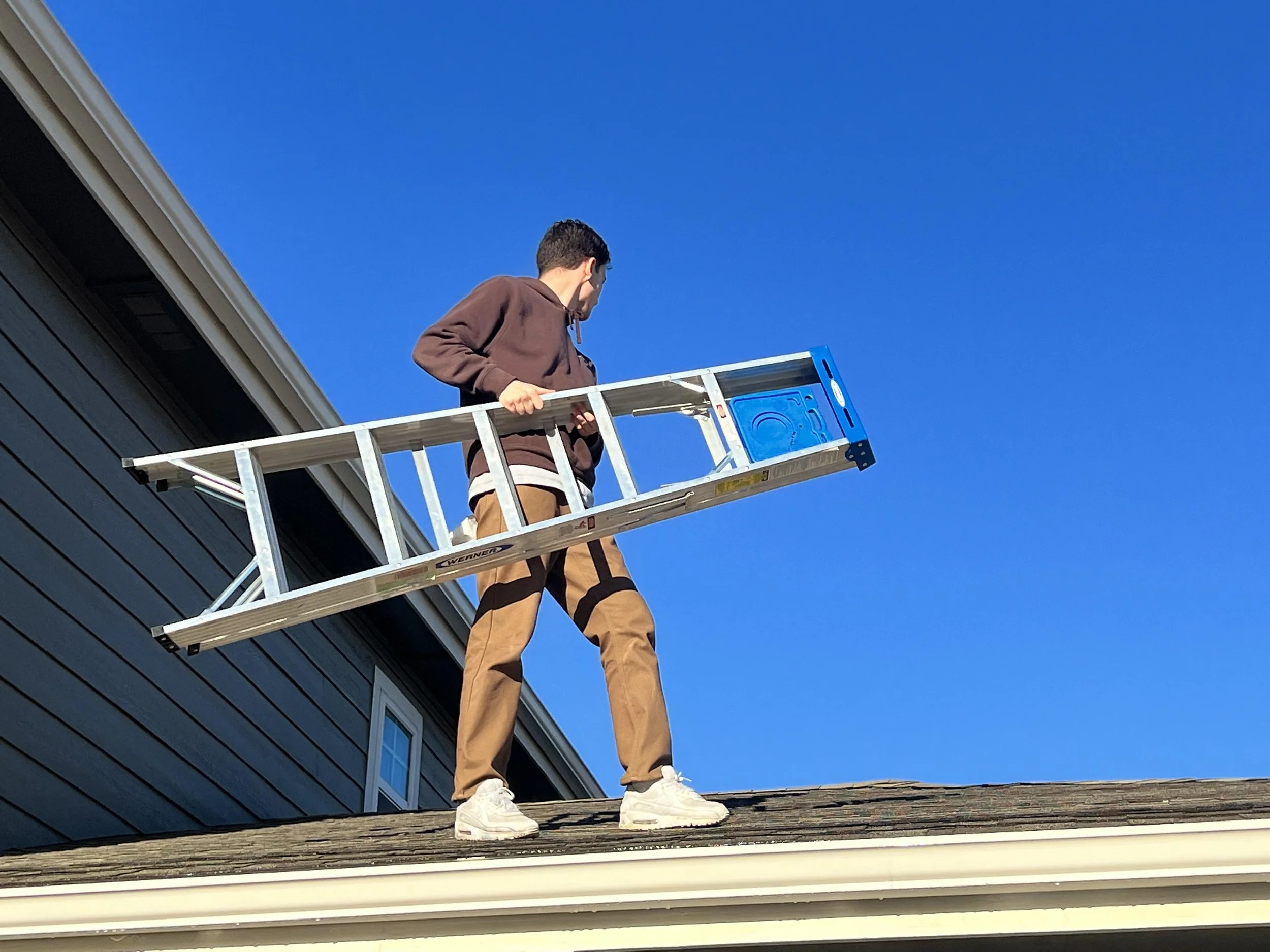 A person on a ladder working on the roof of a house with blue siding and gray shingles.