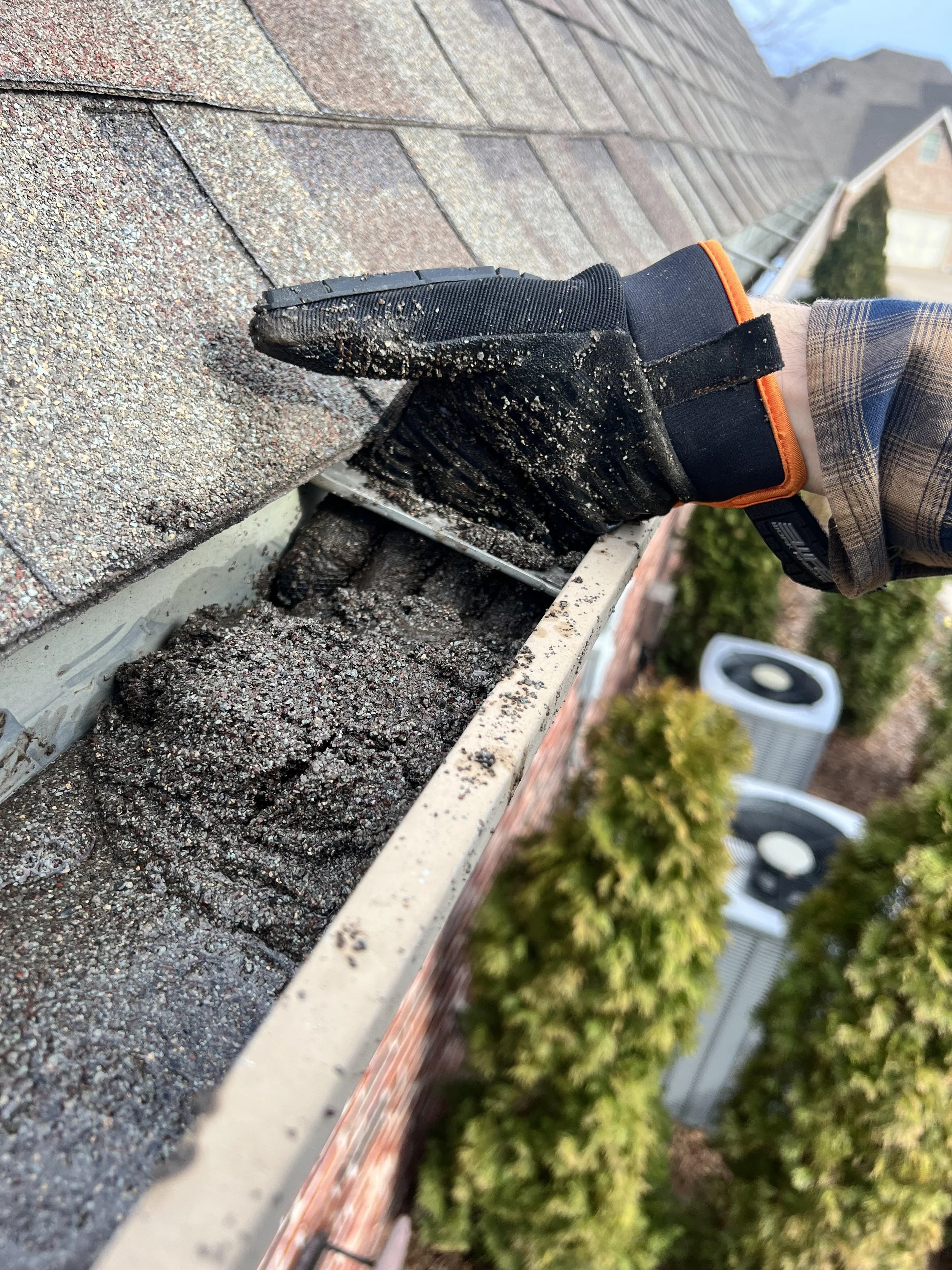Close-up of a gloved hand working on a roof gutter with shingles and house plants in the background.