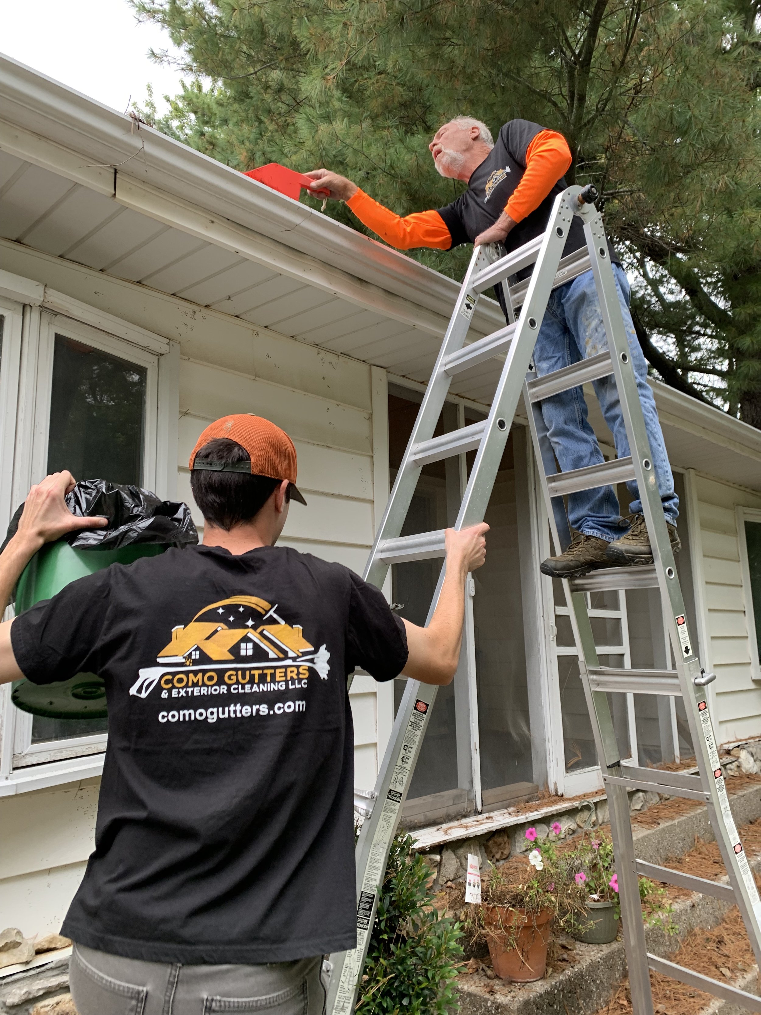 A man on a ladder handing out a red object to another man who is on the ground holding a garbage bag, outside a house with a flower pot and pine trees in the background.