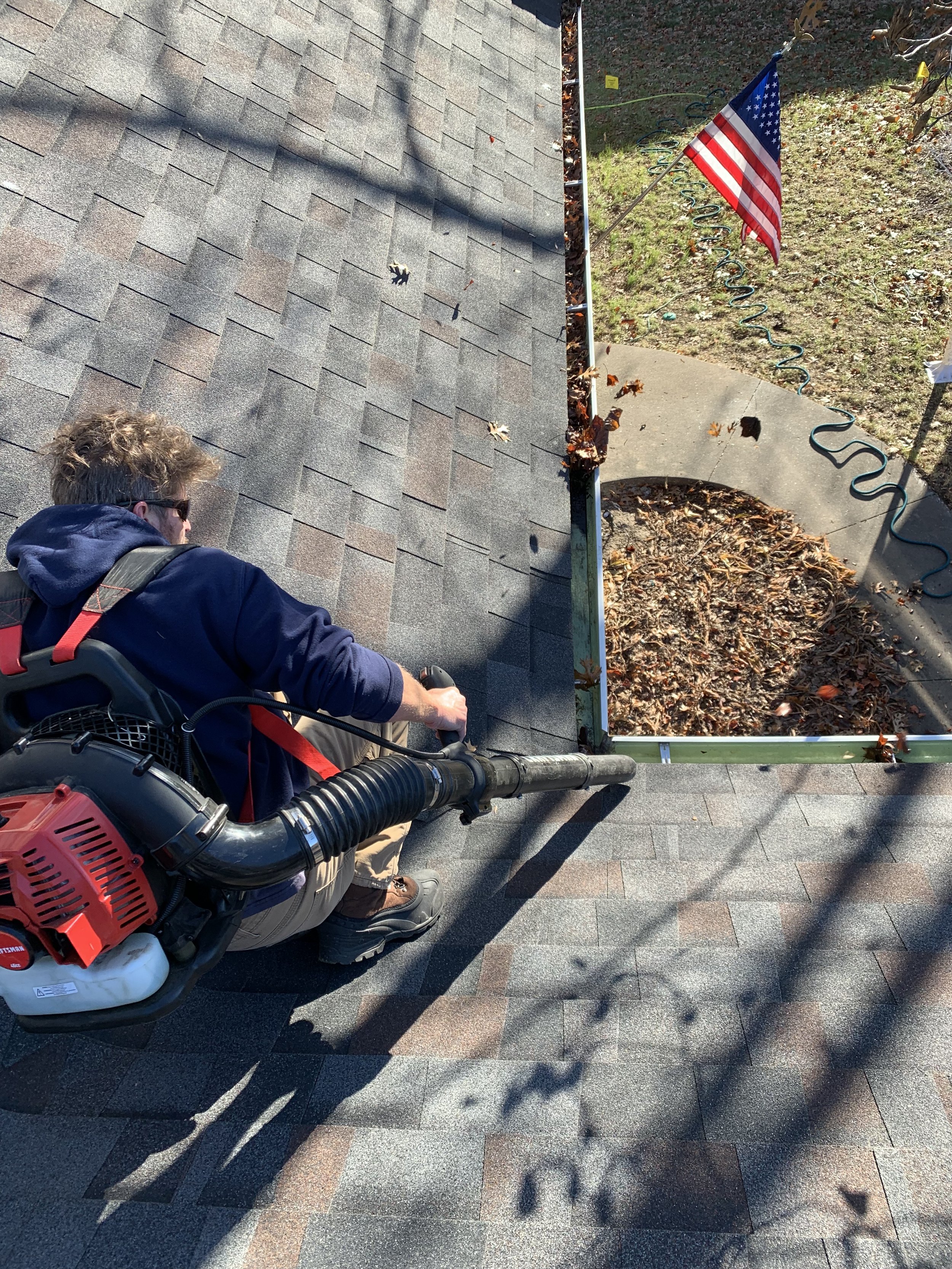 A person in work gear uses a leaf blower to clear leaves from a rooftop with asphalt shingles. An American flag is visible in the background, along with a garden and a sidewalk.