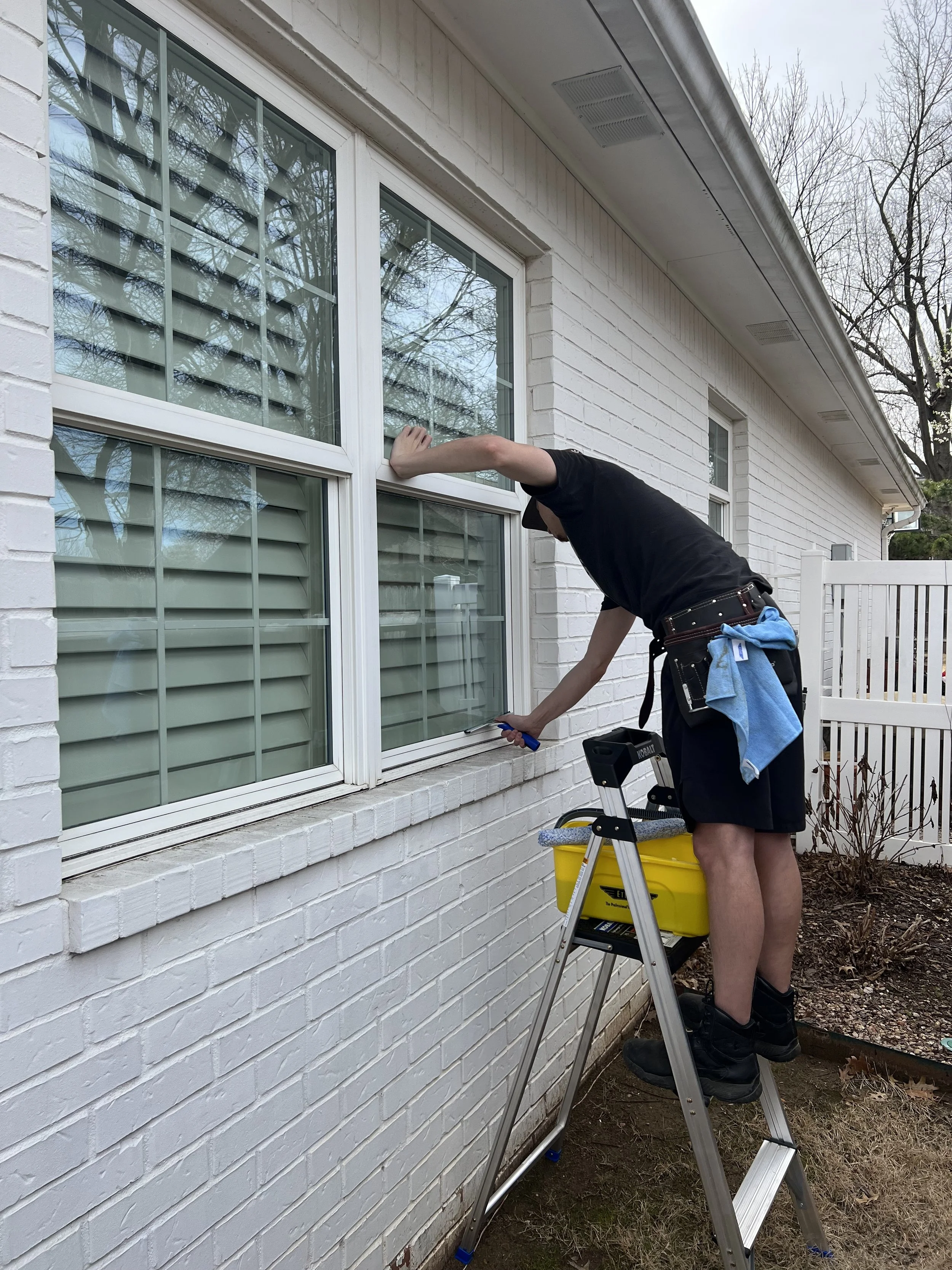 A person wearing black boots, a black cap, a black shirt, and black shorts stands on a step ladder cleaning the exterior window of a white brick house. The person has a towel tucked into their belt, and a yellow bucket with cleaning supplies is placed on the ladder's platform. The house has white shutters, and the yard has some small bushes and leafless trees.