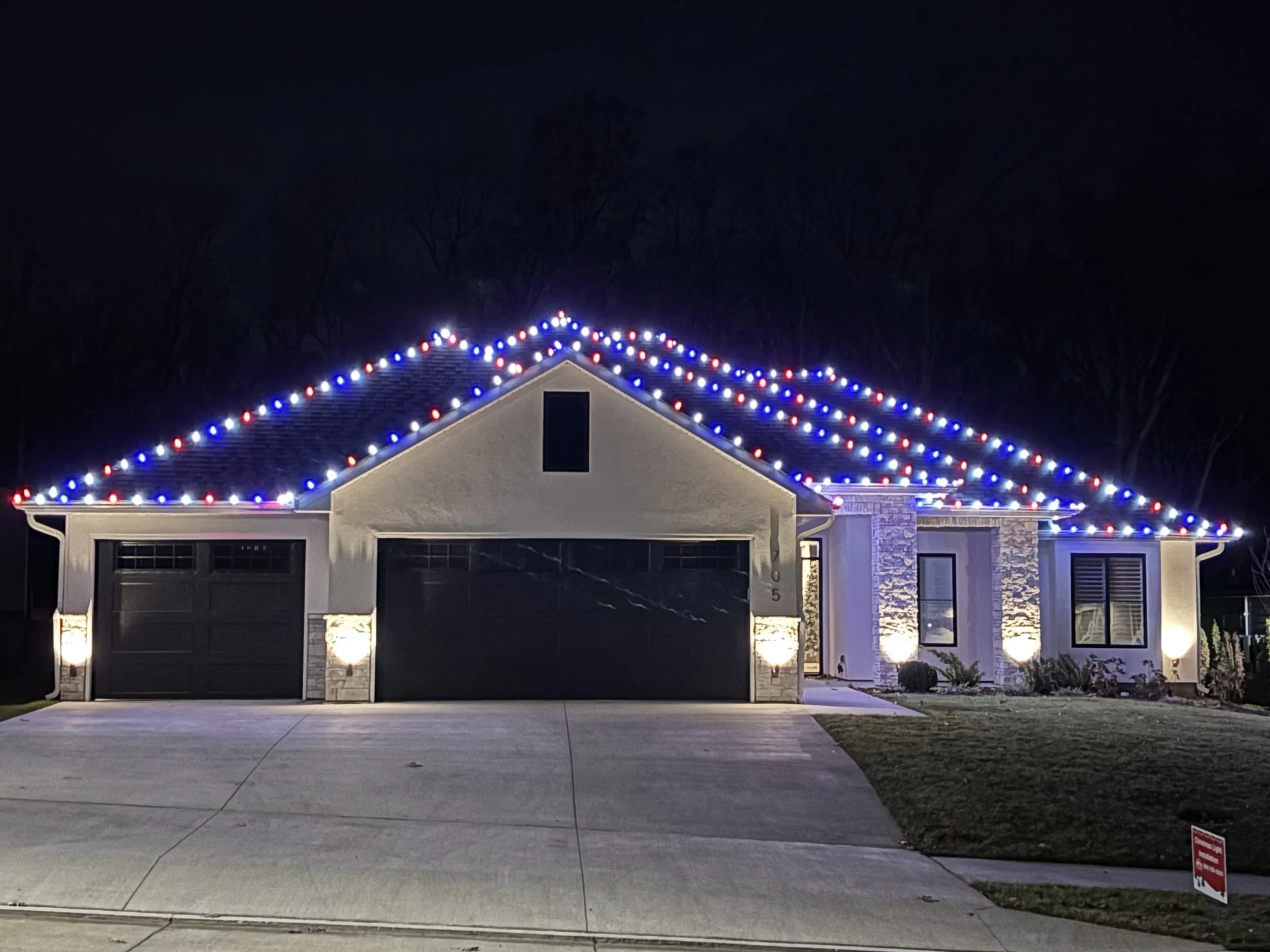 A house decorated with red, white, and blue string lights on the roof at night. The house has a garage, a stone chimney, and a small front yard with a sidewalk, with trees and a dark sky in the background.