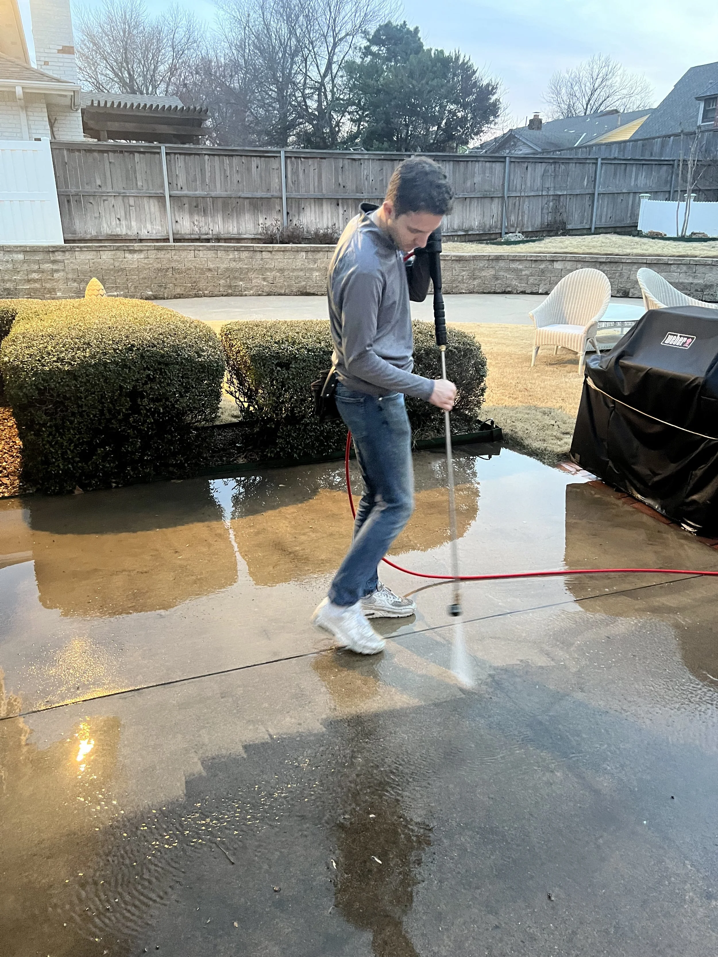 A young man is cleaning a wet concrete driveway with a power scrubber. There are bushes, patio furniture, and a wooden fence in the background.