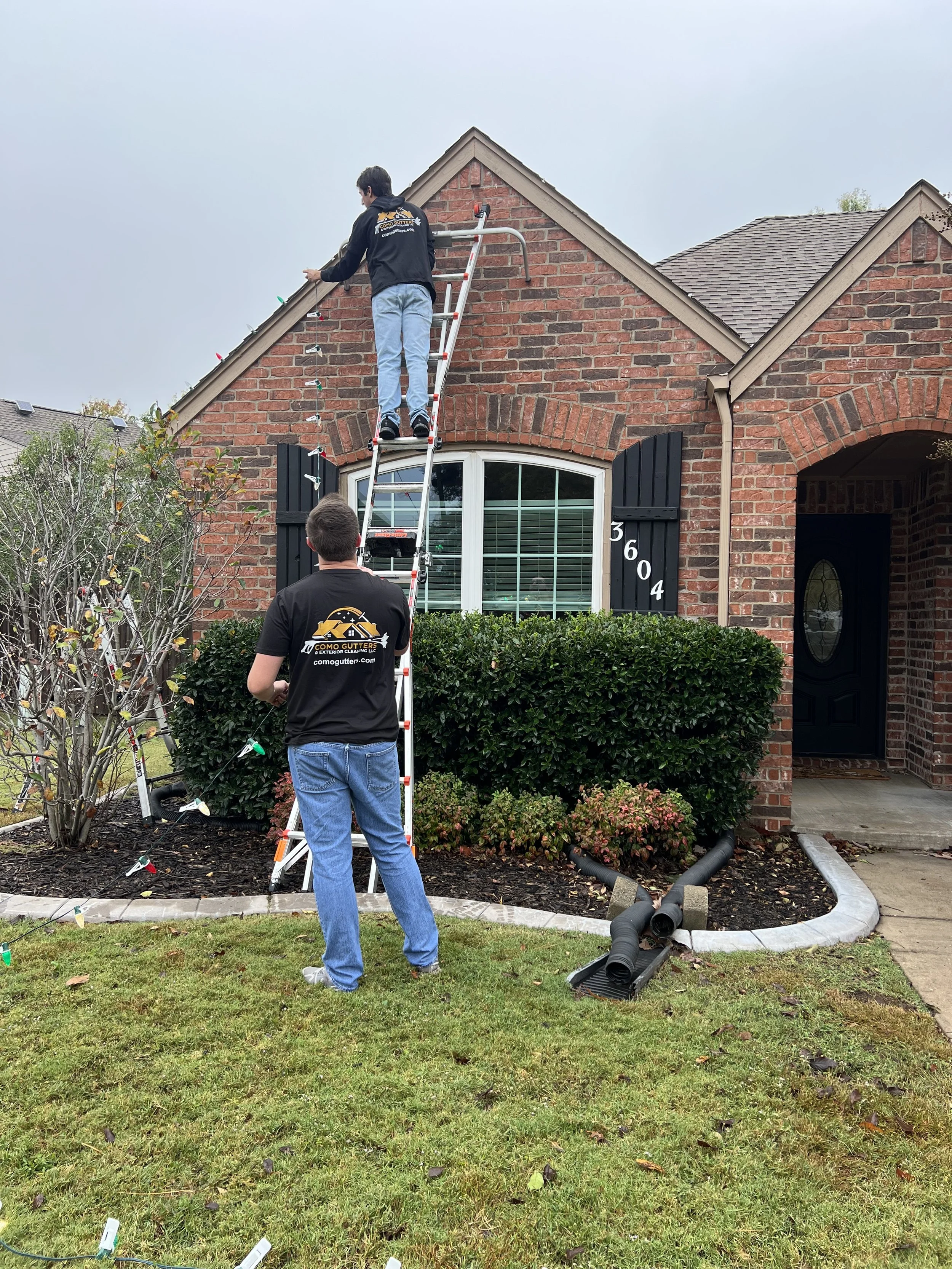 Two workers installing gutter guards on the front of a brick house. One worker on a ladder and the other on the ground, with gutter guard materials and tools nearby.