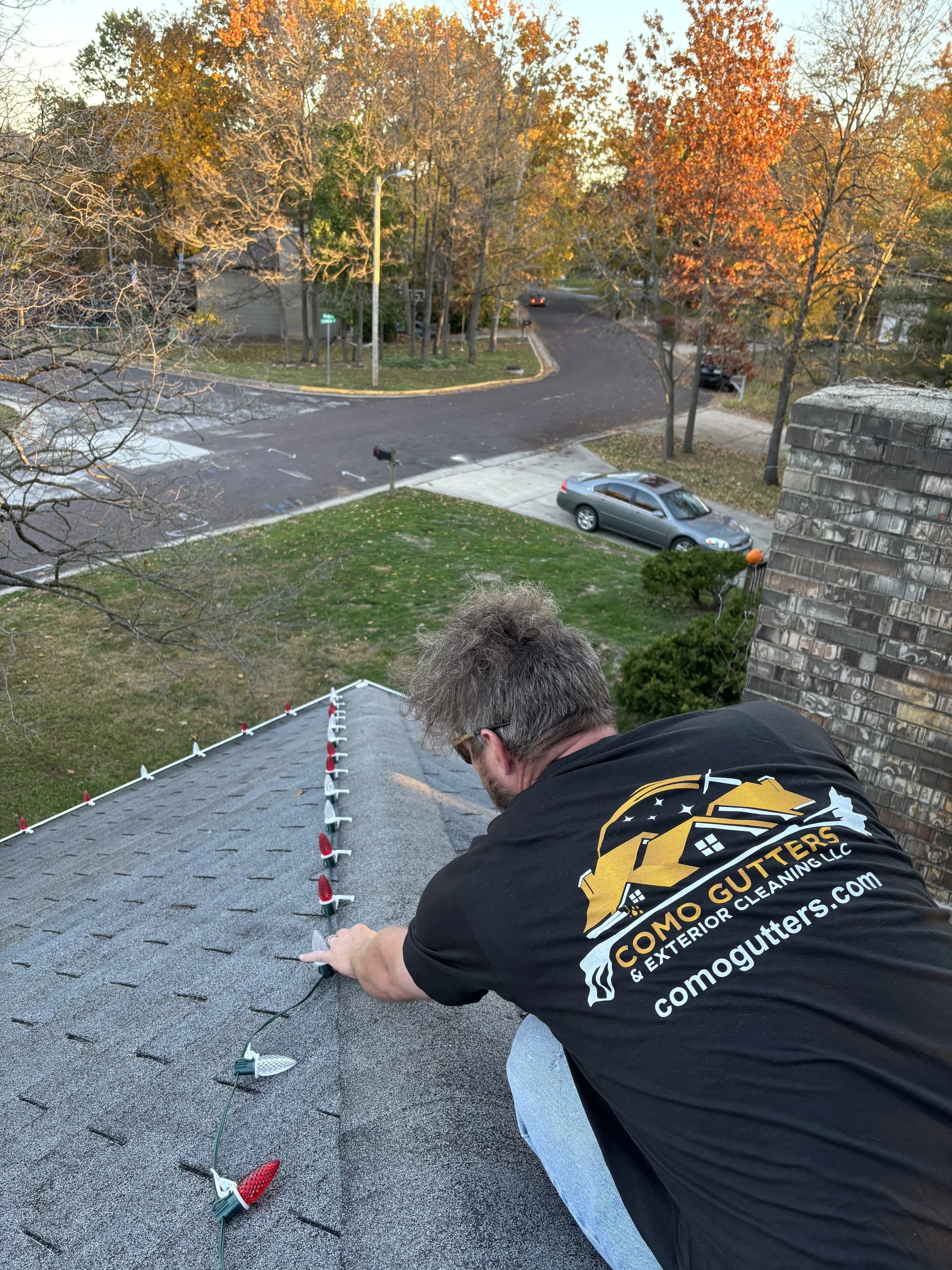 A man installing holiday solar-powered pathway lights on a black shingled roof, with a suburban street scene with trees with autumn foliage and a parked car in the background.