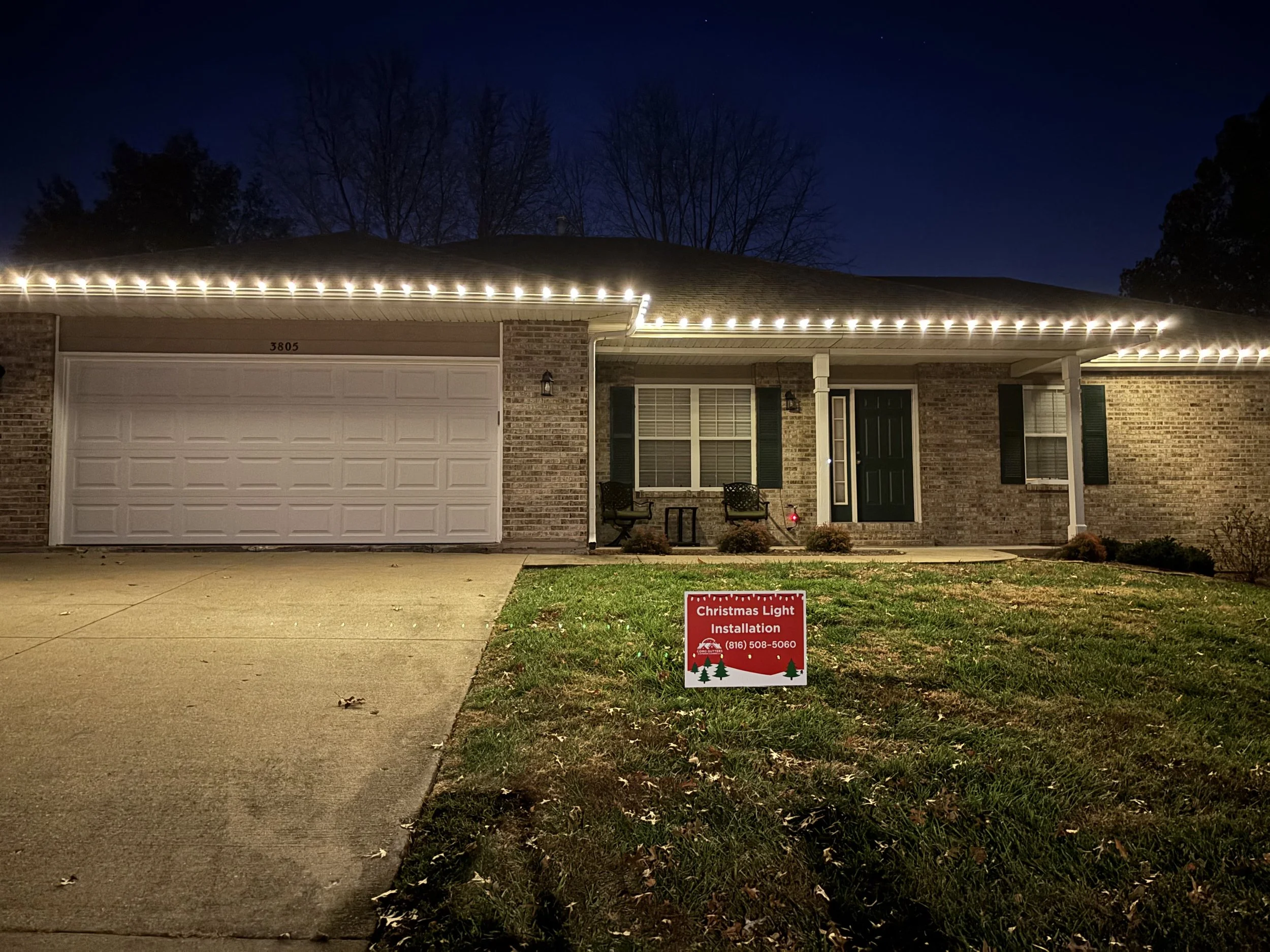 Front view of a brick house with a white garage door and porch, illuminated Christmas lights outline the roof, a red holiday sign on the lawn reads 'Christmas Light Installation' with contact number and Christmas tree images, during nighttime with a dark sky and leafless trees in the background.