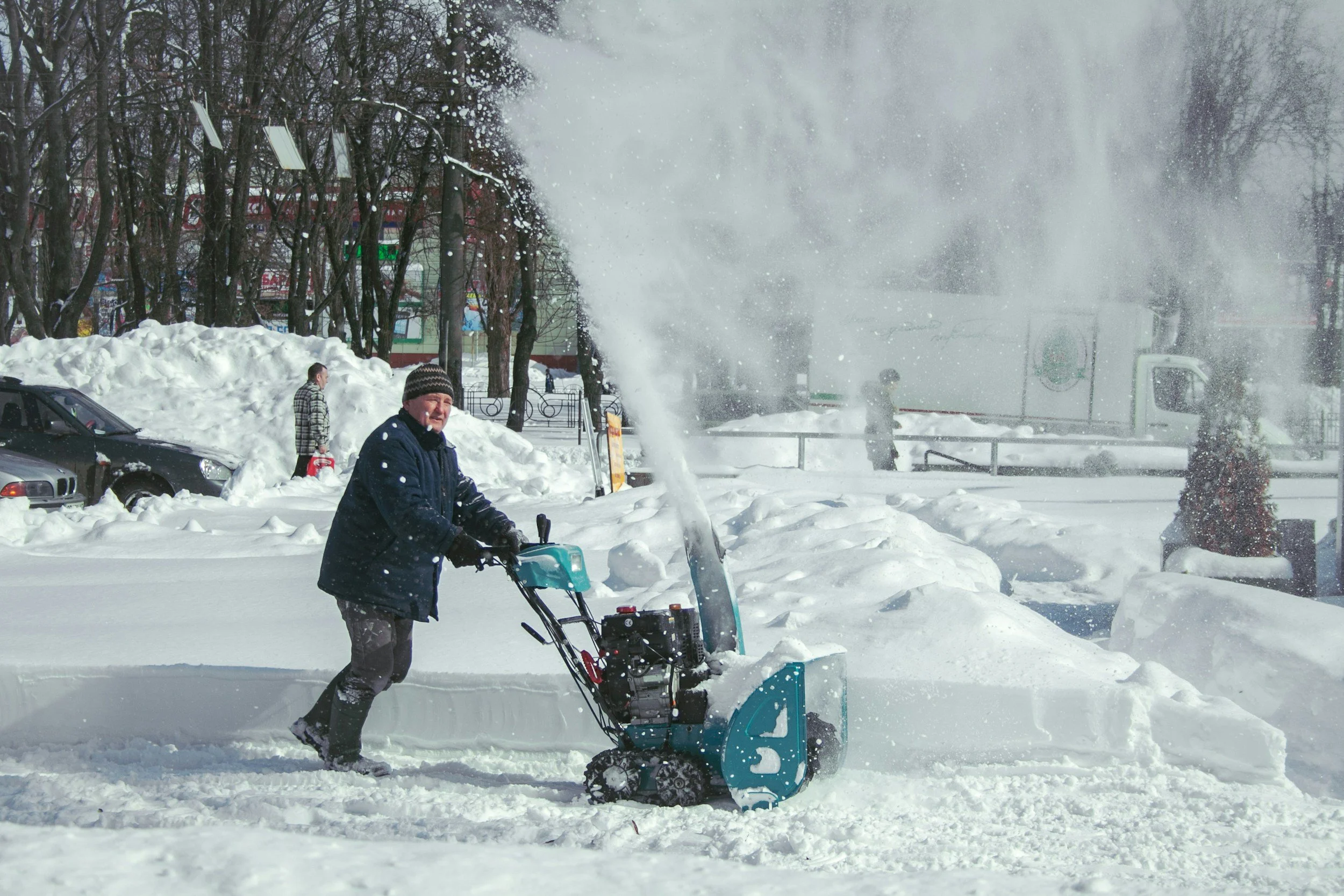 A man wearing a black jacket and a knit hat is operating a snow blower on a snow-covered sidewalk, with large snow piles behind him. In the background, a woman is snow blowing near a fence, and a man is walking along the cleared path. There are leafless trees, parked cars, and a building with signs in the distance. Snow is being blown into the air as the snow blower operates.