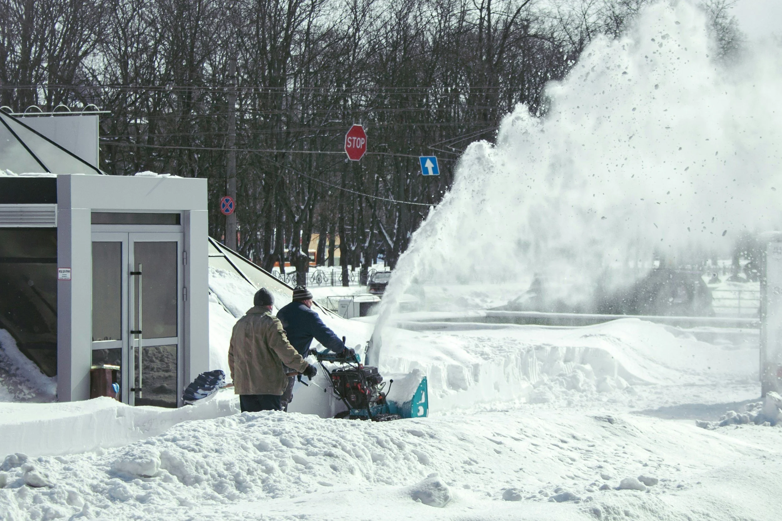 Two people are clearing snow from a sidewalk with snow blowers, with a large snow drift falling on a window and a stop sign in the background.