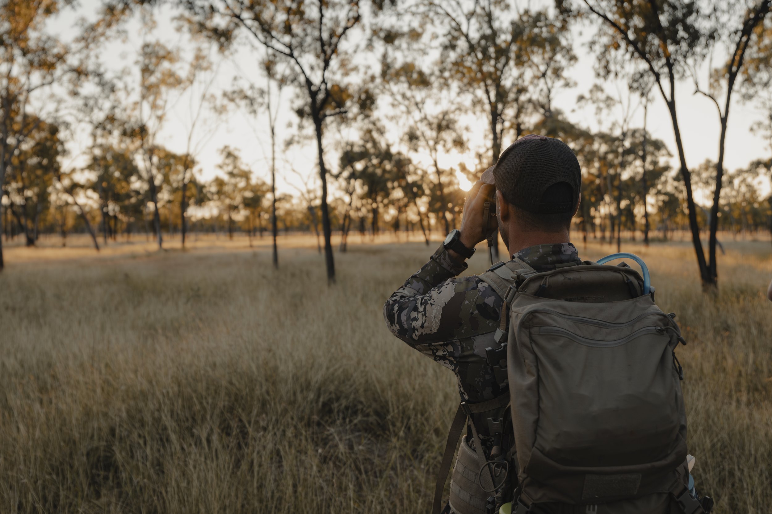 A person in outdoor camouflage clothing and a cap is standing in a field with tall grass, taking a photo of a wooded area during sunset or sunrise.