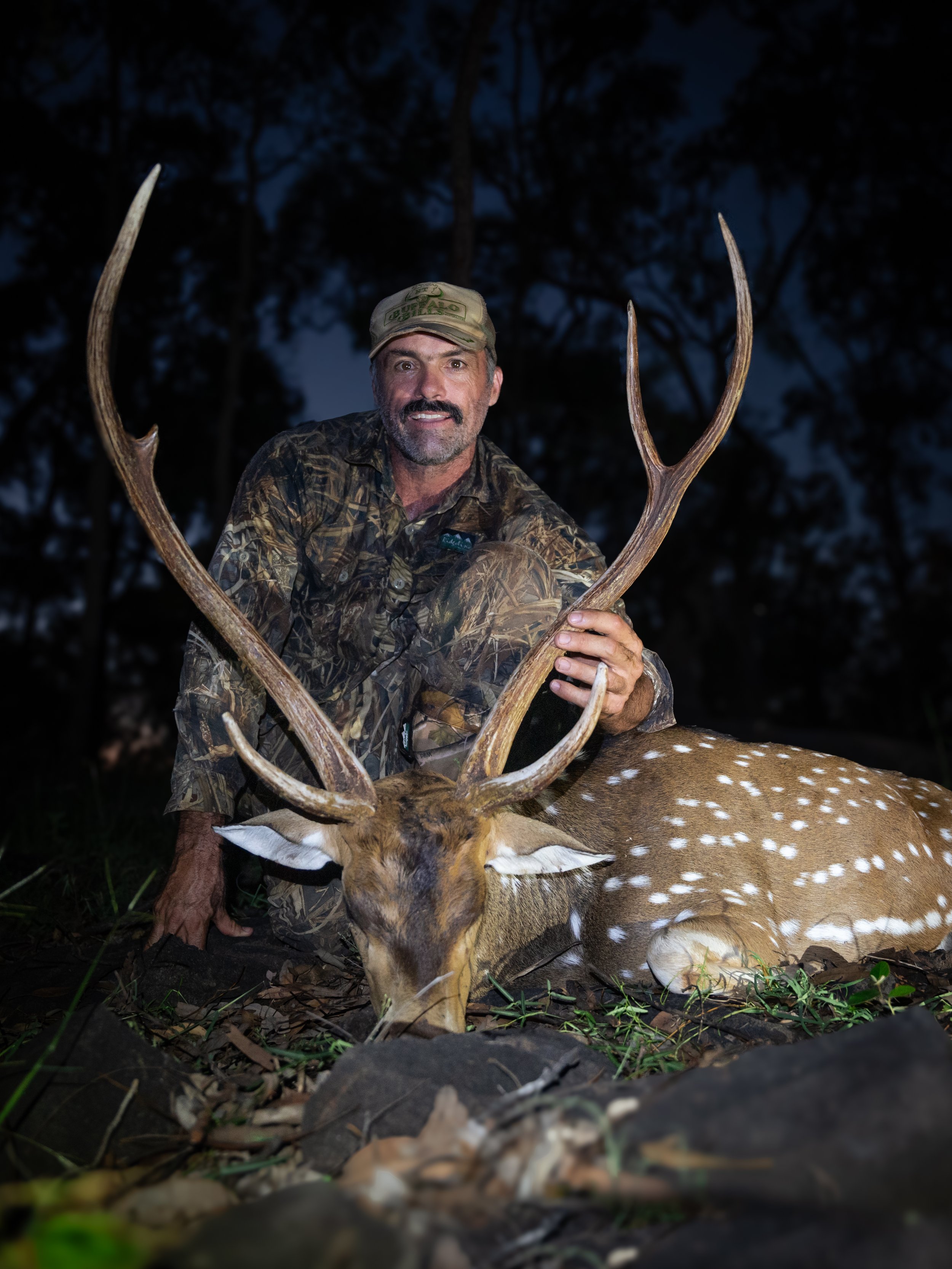 A man in camouflage clothing holding a large deer with antlers at night outdoors.