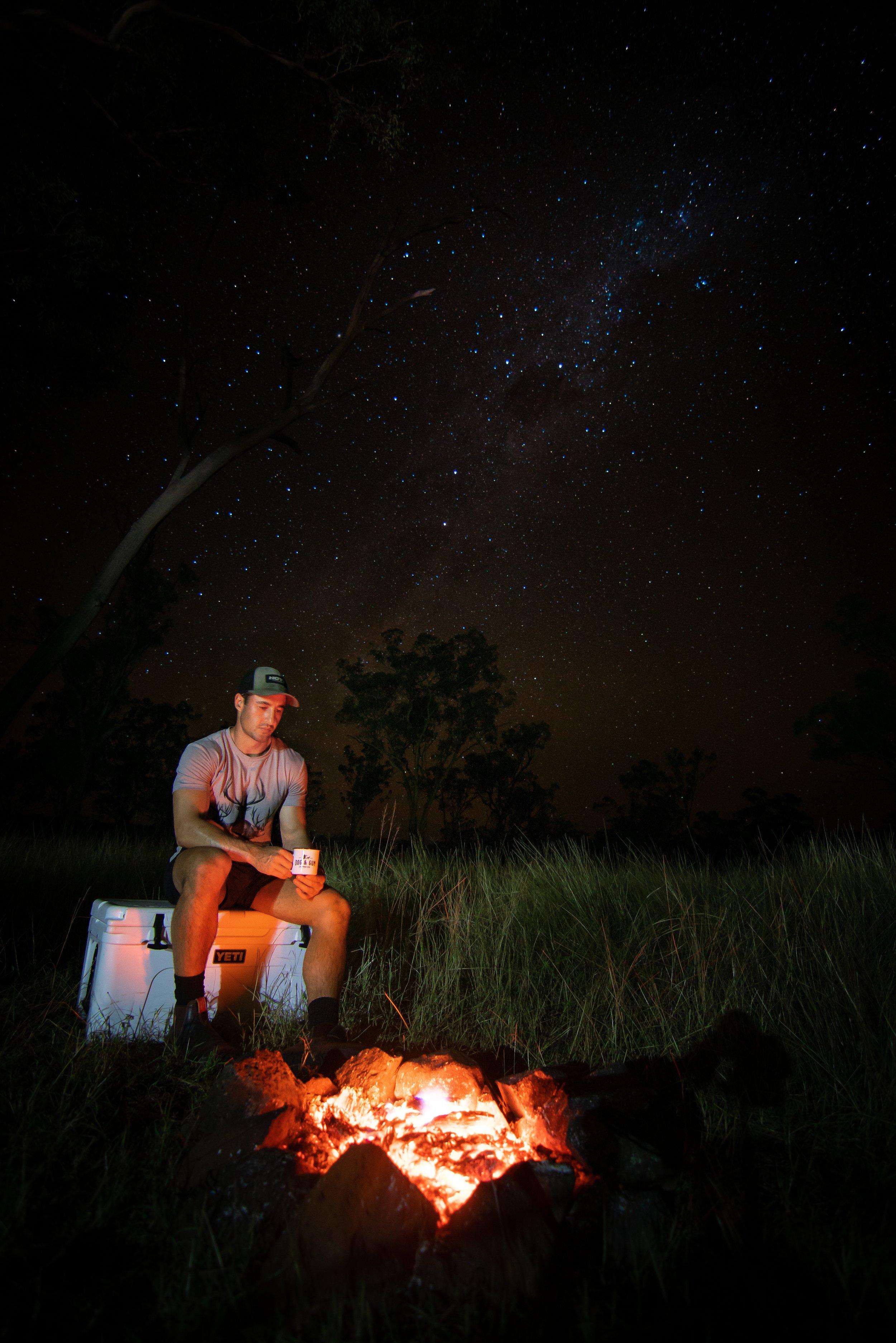 A man sitting on a cooler by a campfire at night, holding a mug and looking down, with a star-filled sky and trees in the background.