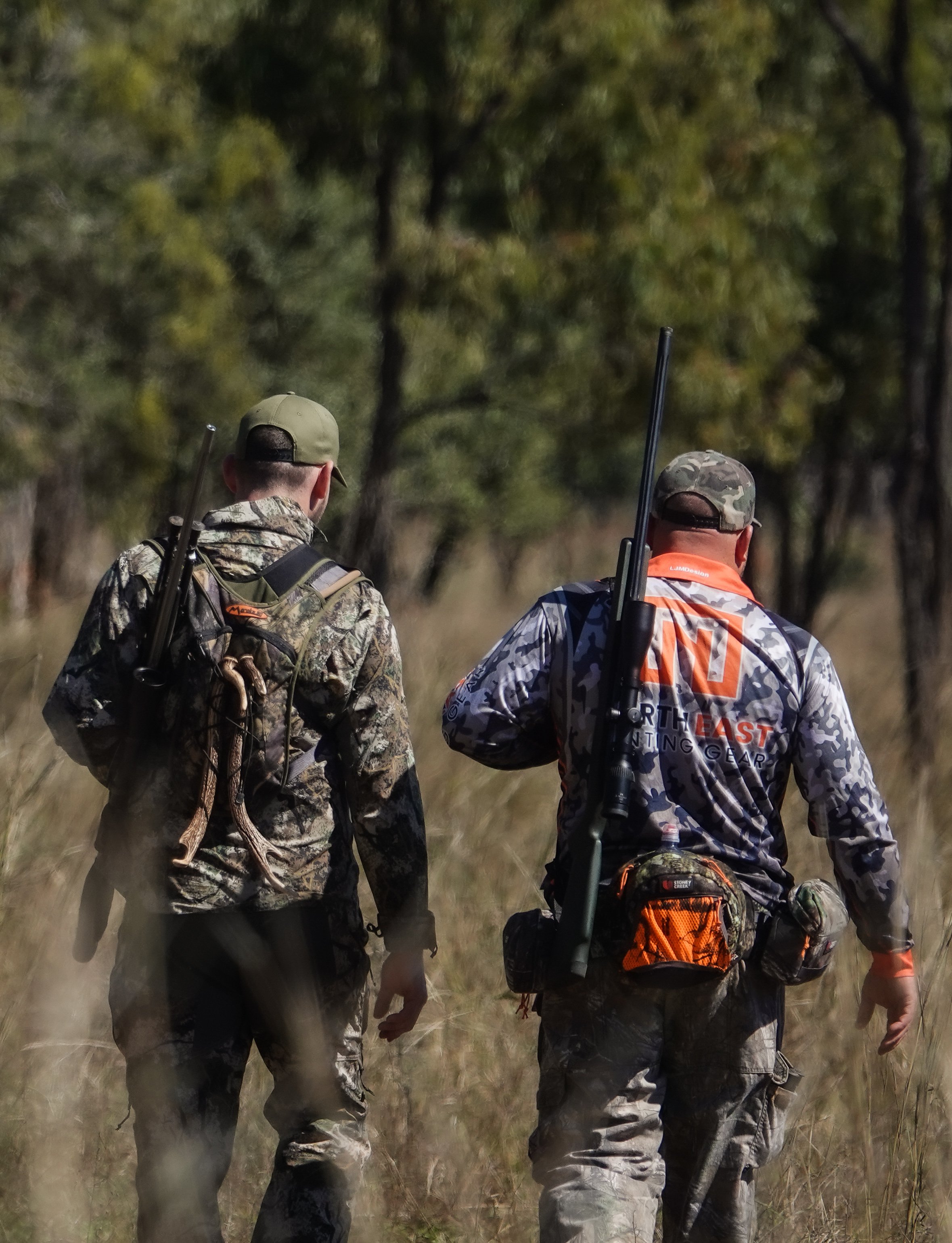 Two men dressed in camouflage hunting gear walking through a grassy field with trees in the background, carrying rifles.