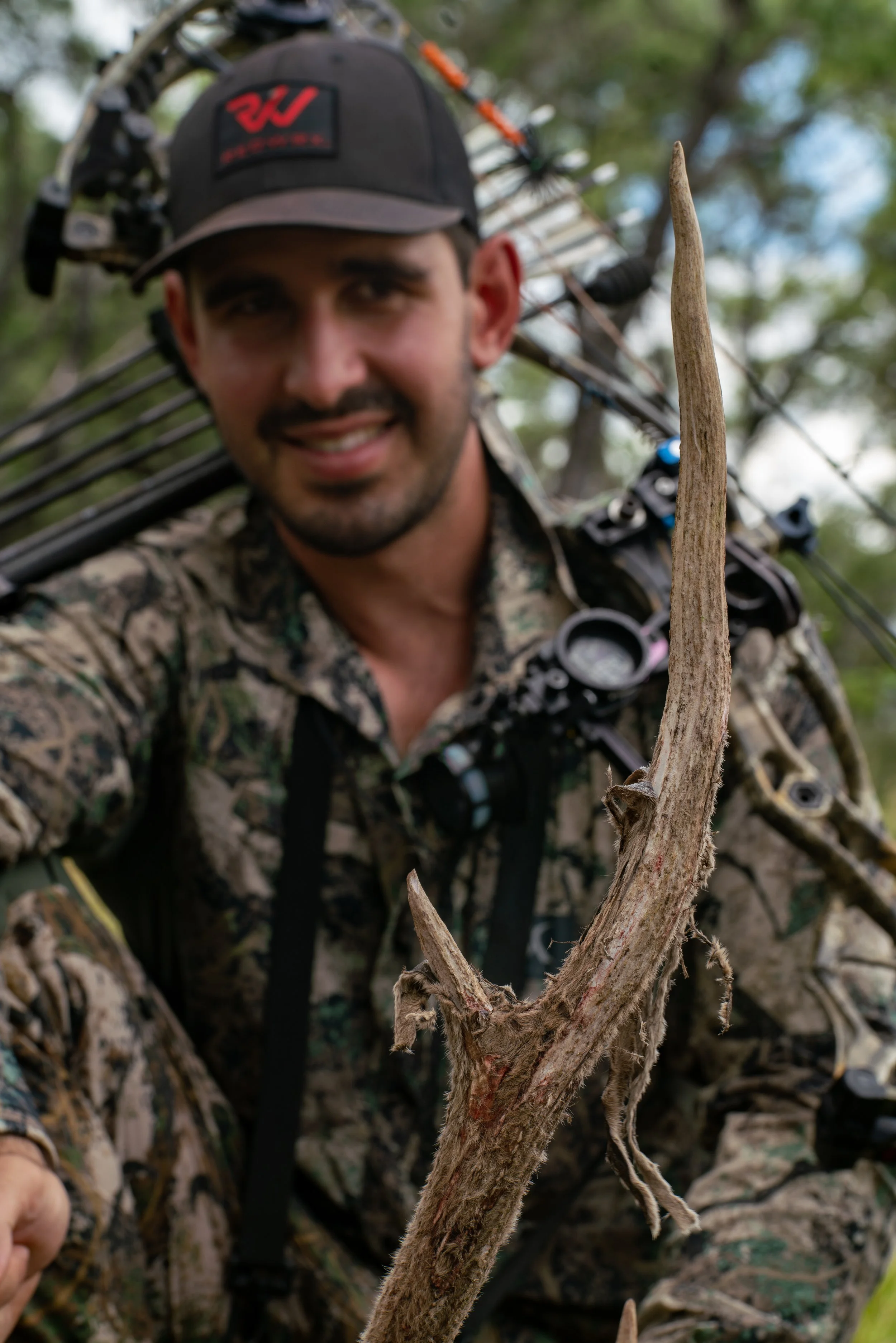 A man in camouflage clothing and a baseball cap holding a bow, standing outdoors among trees, with a dry branch in the foreground.