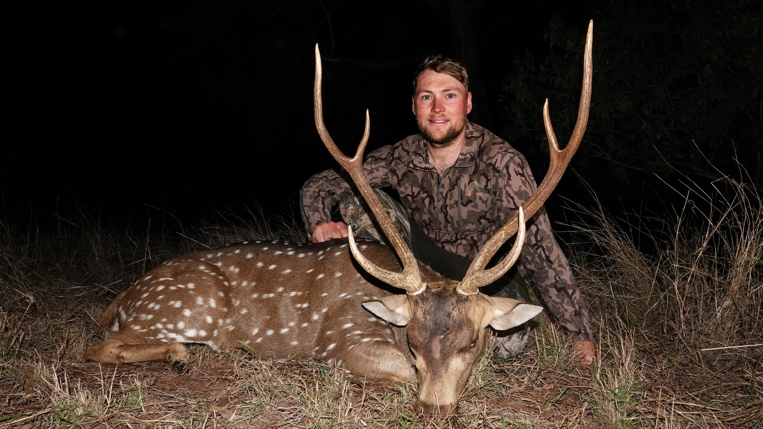 A man in camouflage clothing kneels behind a large deer he has hunted, with antlers, at night outside in tall grass