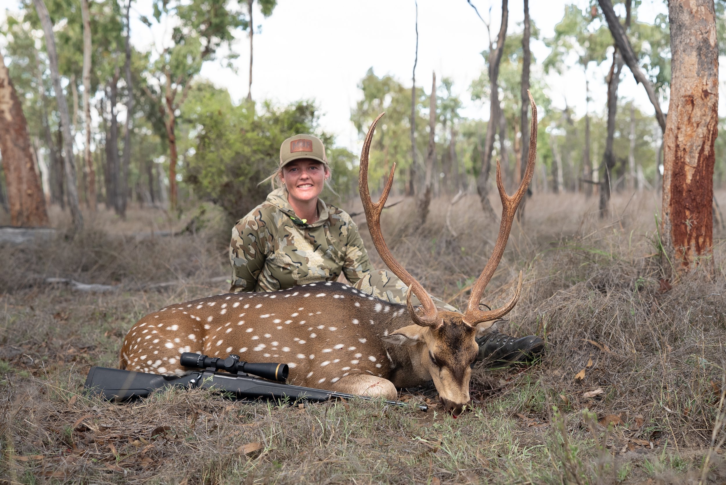 A woman in camouflage clothing and a hat outdoors in a forest, sitting next to a dead deer with antlers, and a rifle resting on the ground.