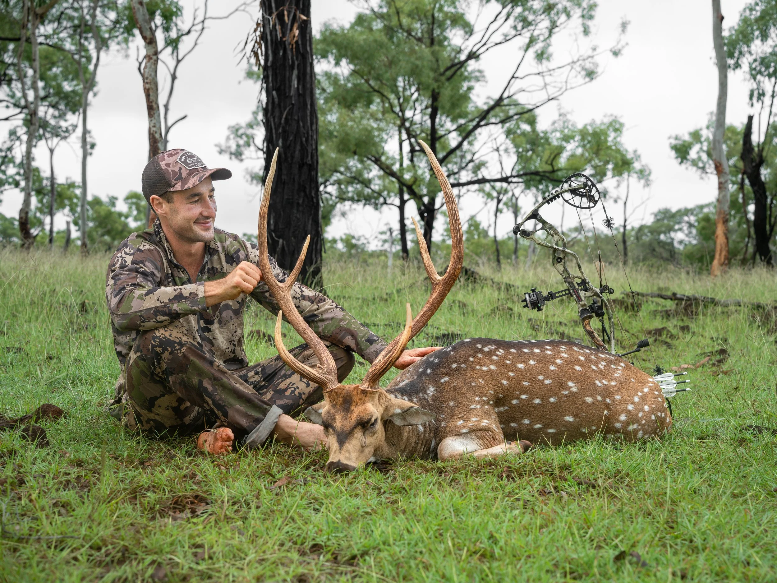 A man dressed in camouflage hunting gear sitting in a forested area, holding antlers of a freshly hunted deer with a bow and arrow nearby, smiling.